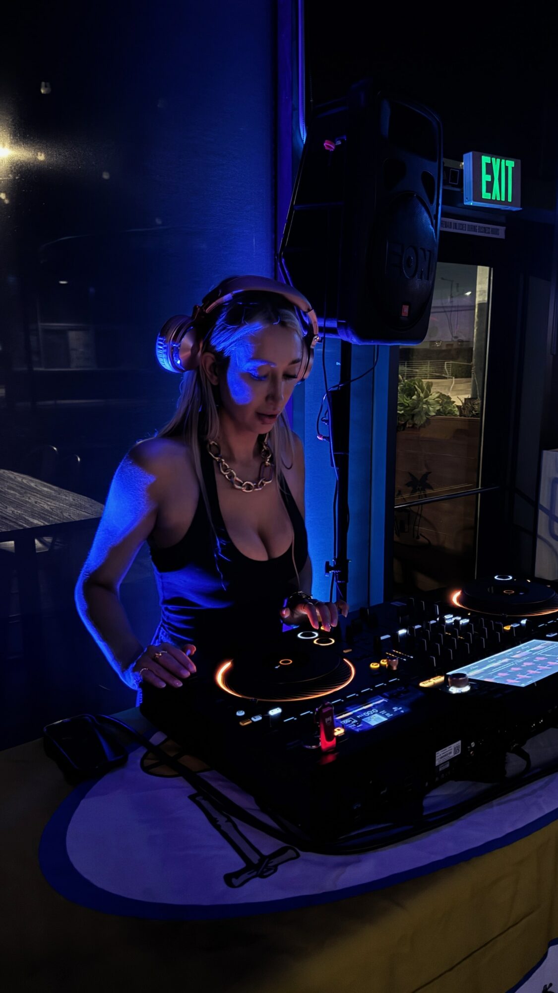 Person DJing at a booth with equipment, illuminated by blue lighting, in a dark indoor setting.