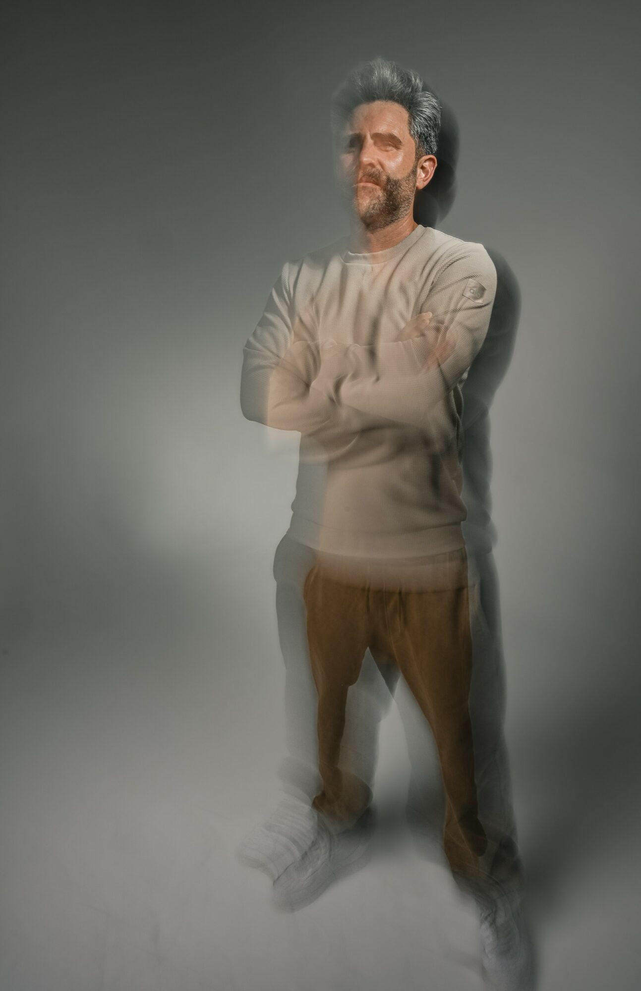 Man with crossed arms, beard, and curly hair stands against a gray background, looking to the side.