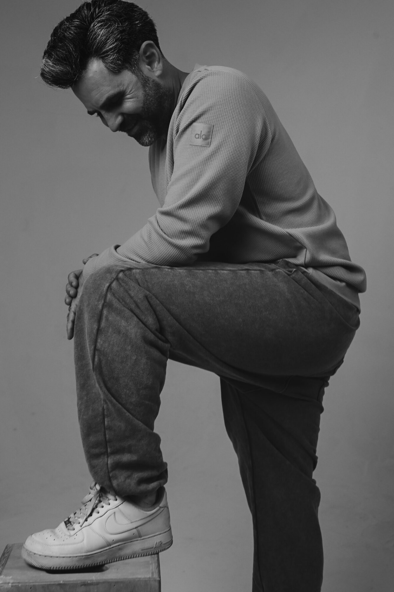 Man with beard and styled hair, wearing casual clothes, balancing on one foot with the other foot on a surface, in black and white