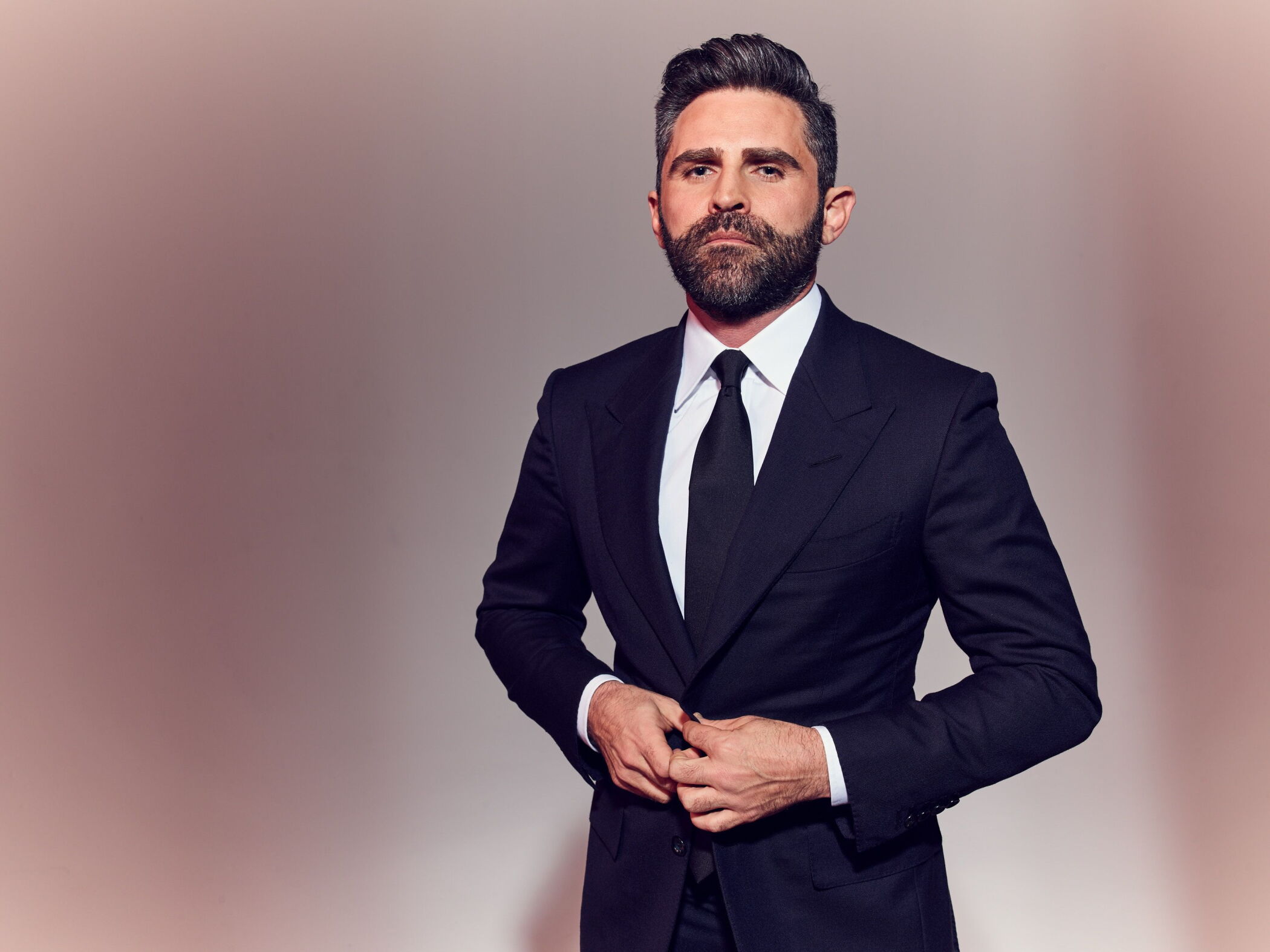 Man in a dark suit and tie, with a beard, standing with hands clasped, against a plain background.