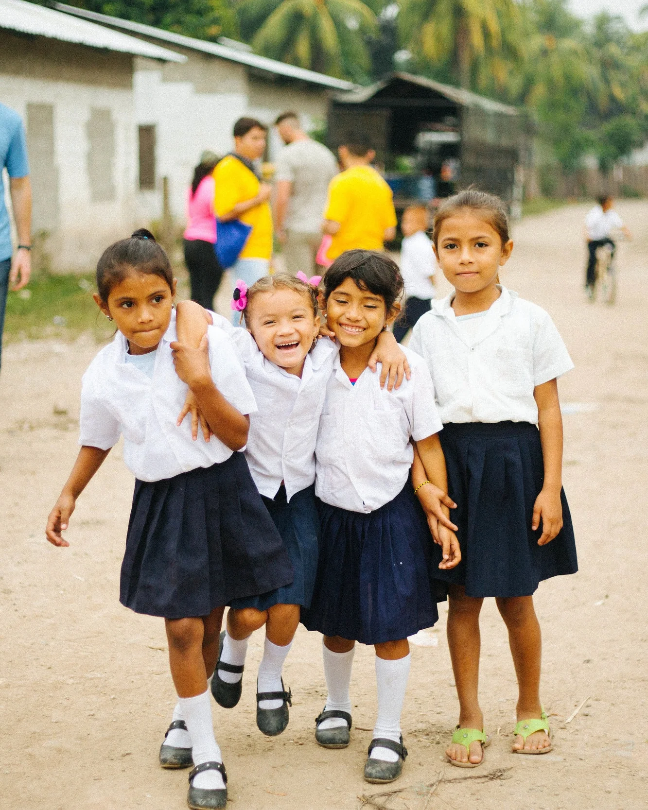 Four children in school uniforms smiling and walking together outdoors, with a group of people and buildings in the background.