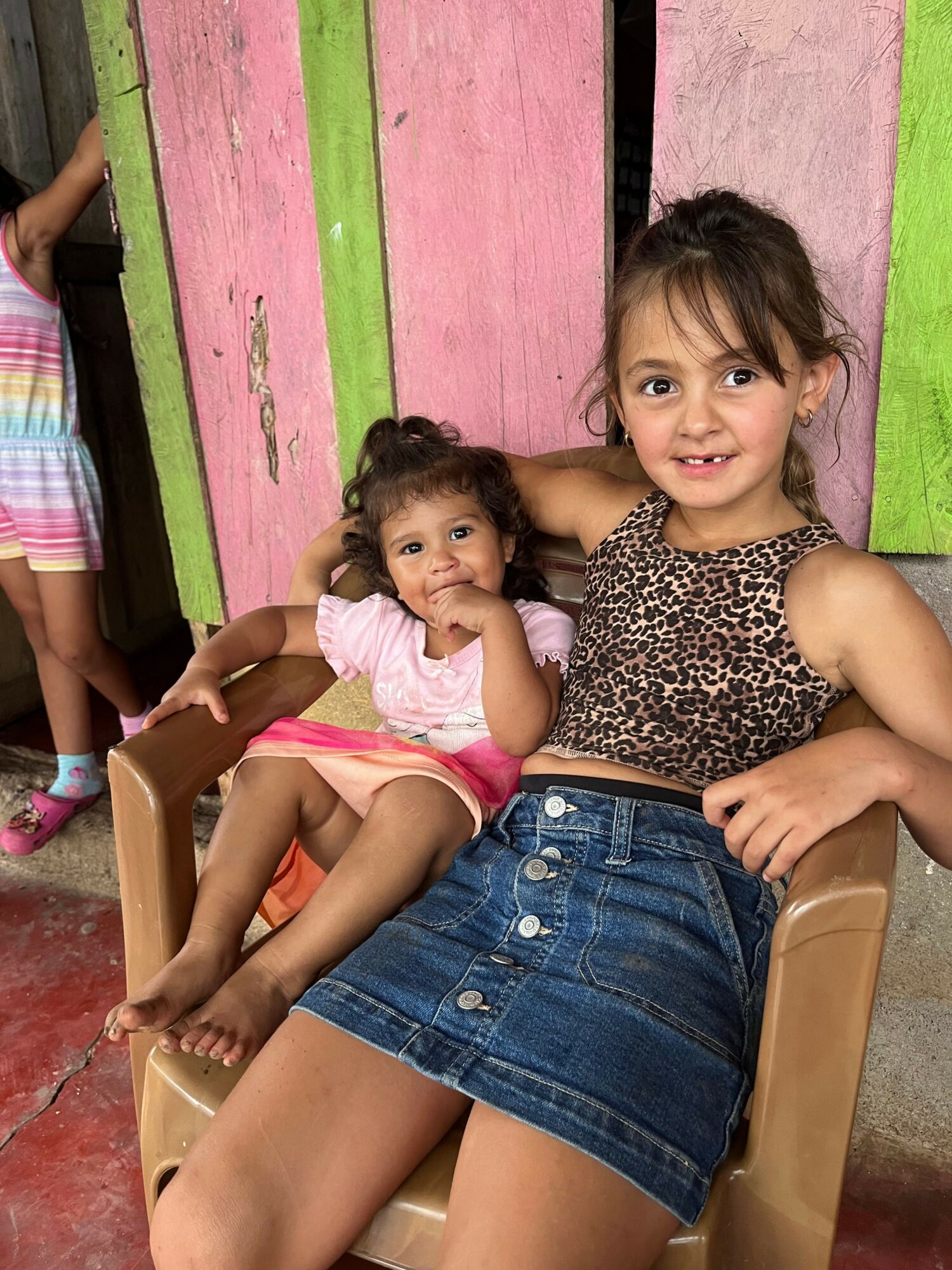 Two young girls sitting on a chair in front of a colorful wooden wall, smiling at the camera.