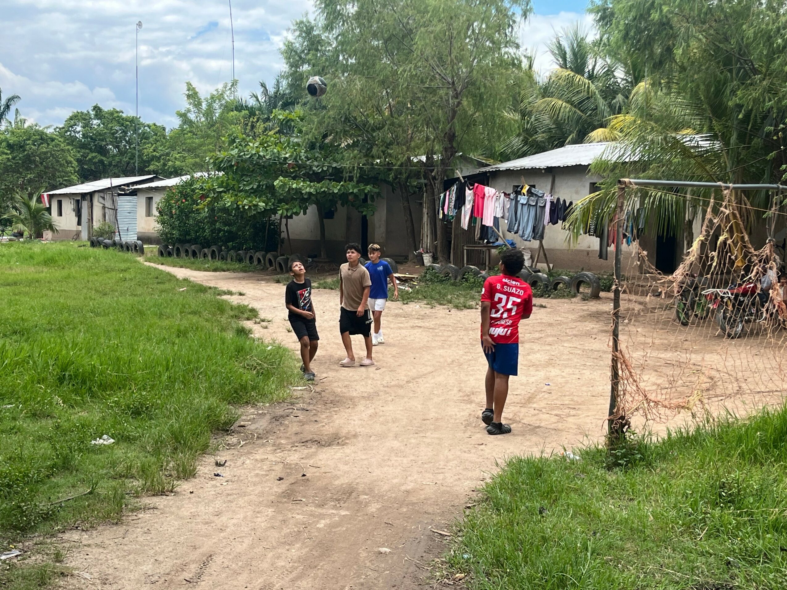 Four people playing soccer on a dirt field with houses and trees in the background.