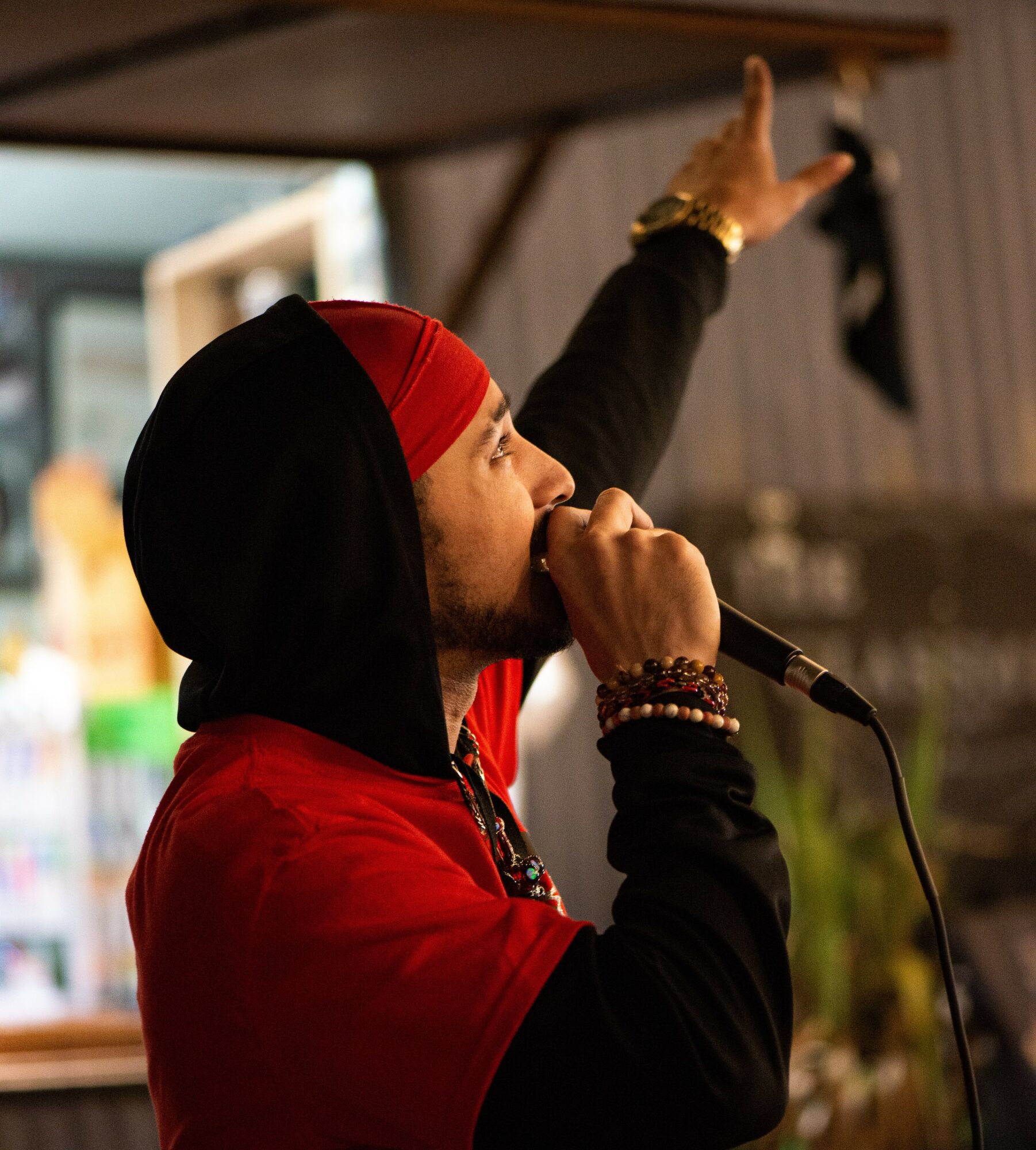 Person singing into a microphone with one arm raised, wearing a red and black head covering and jewelry.