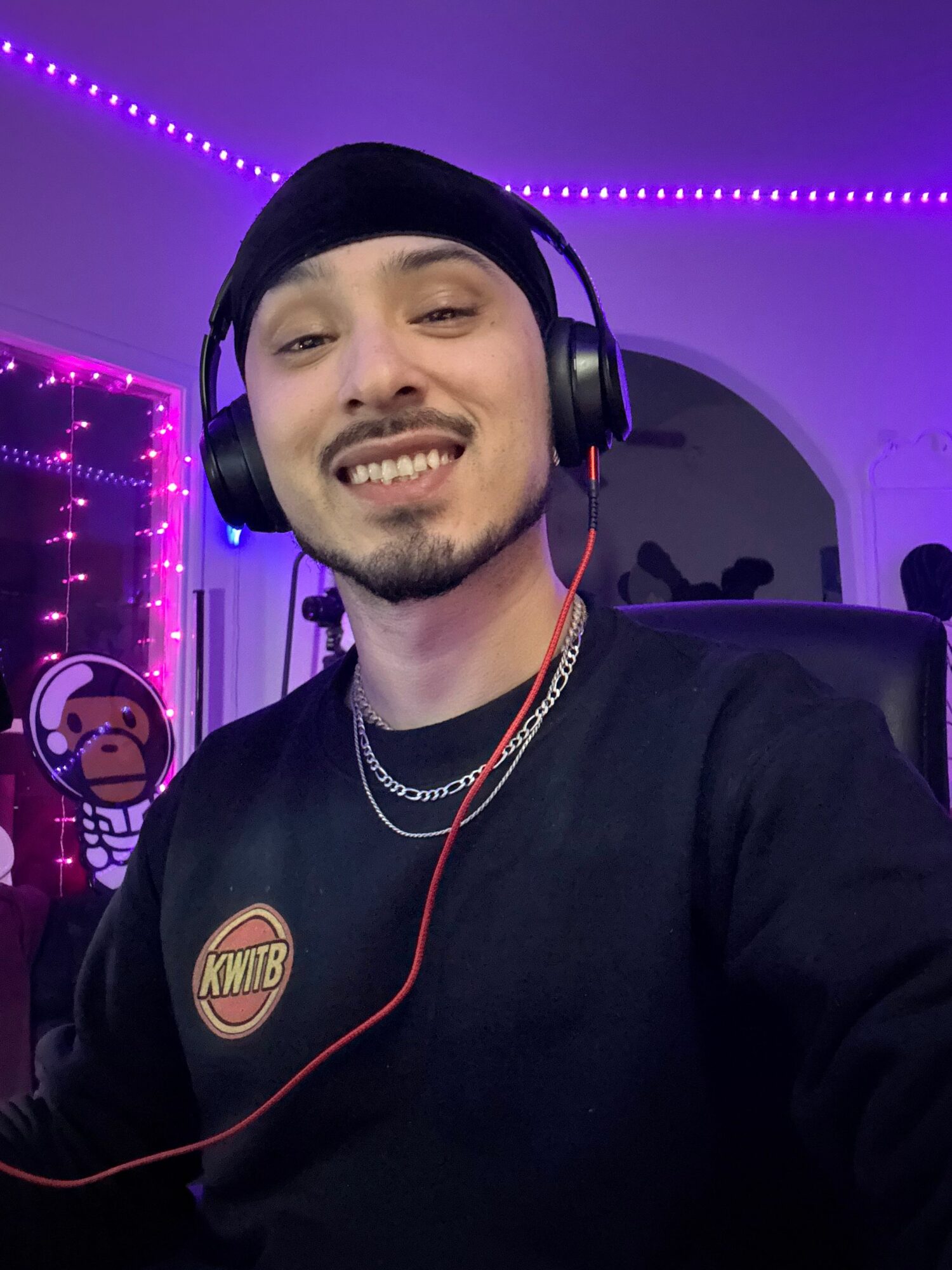 Young man wearing headphones and a black beanie, smiling in a room with purple LED lights, wearing a black shirt with a logo.