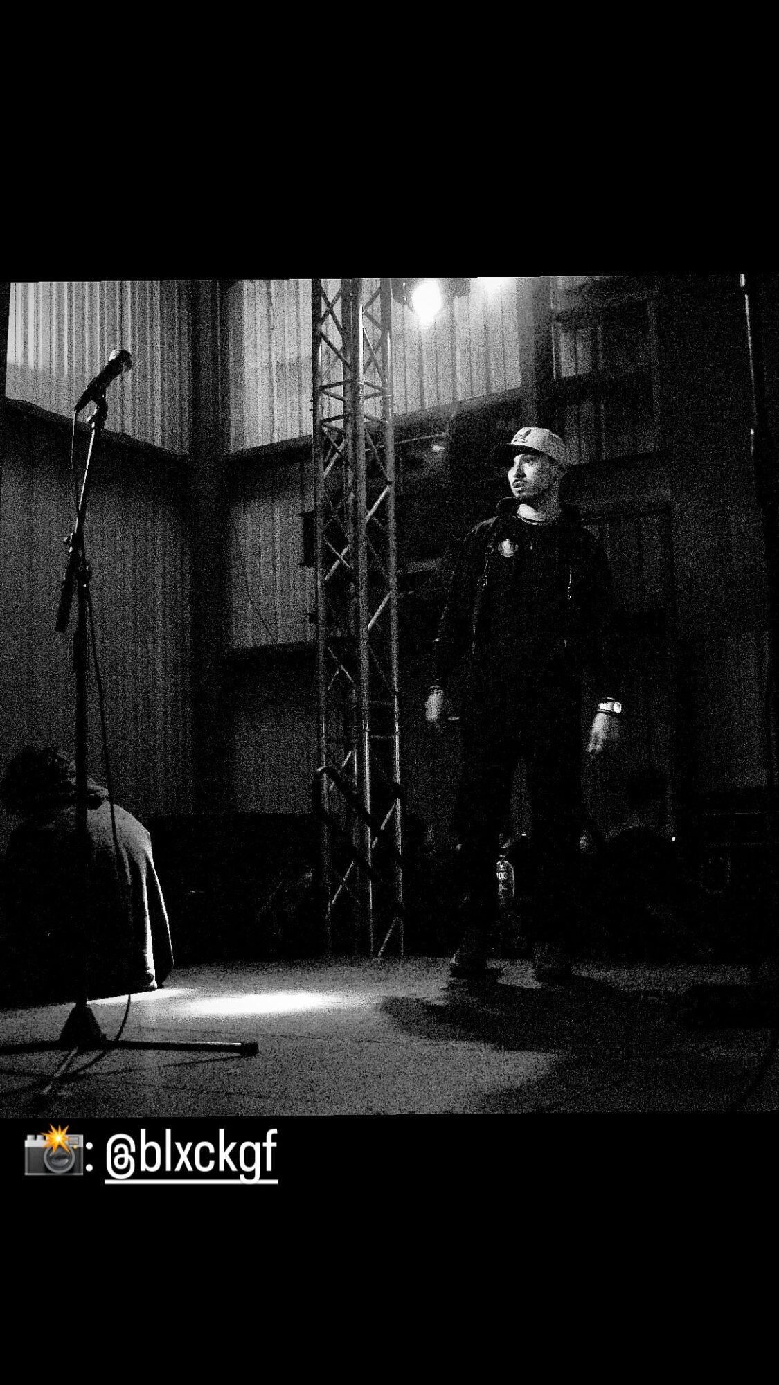 Person standing on stage with microphone, stage equipment, and a person sitting nearby, in a dark indoor setting.