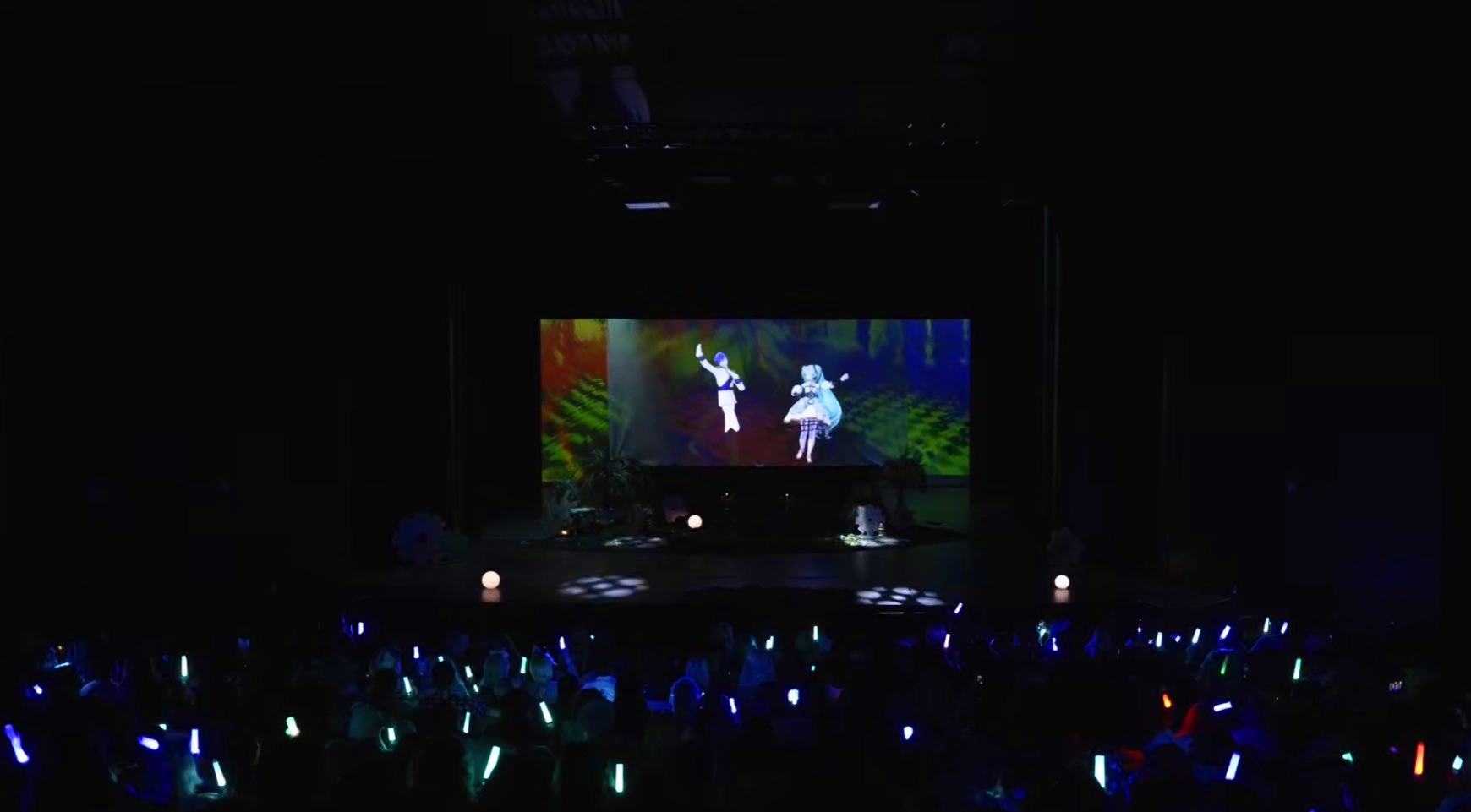 Stage with animated characters projected on screen, audience holding colorful glow sticks, dark background.