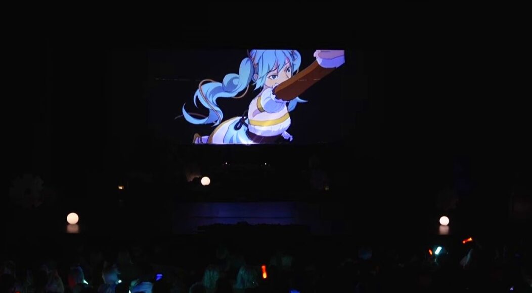 A large screen displays an animated character with blue hair and a white outfit, in a dark room with audience silhouettes.