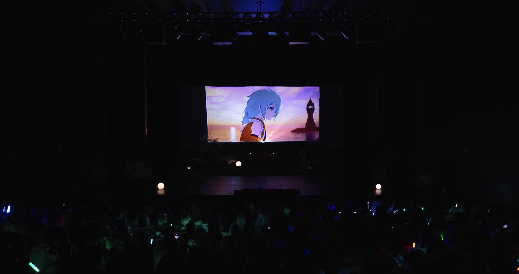 A dark theater with a large screen displaying an animated character with blue hair, and an audience in front.