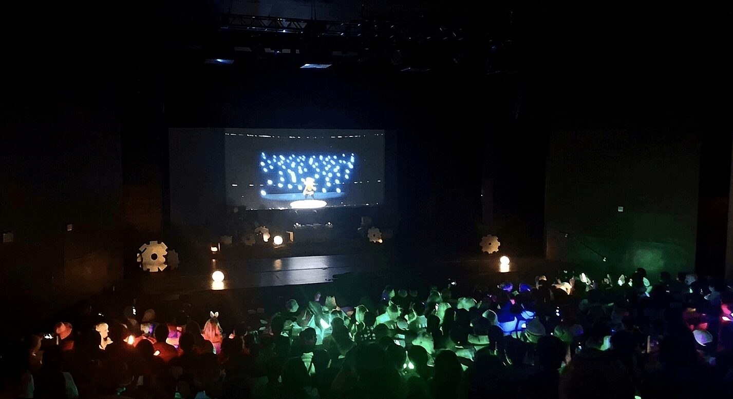 Dark theater with a stage, screen displaying blue visuals, and audience illuminated by colorful lights.