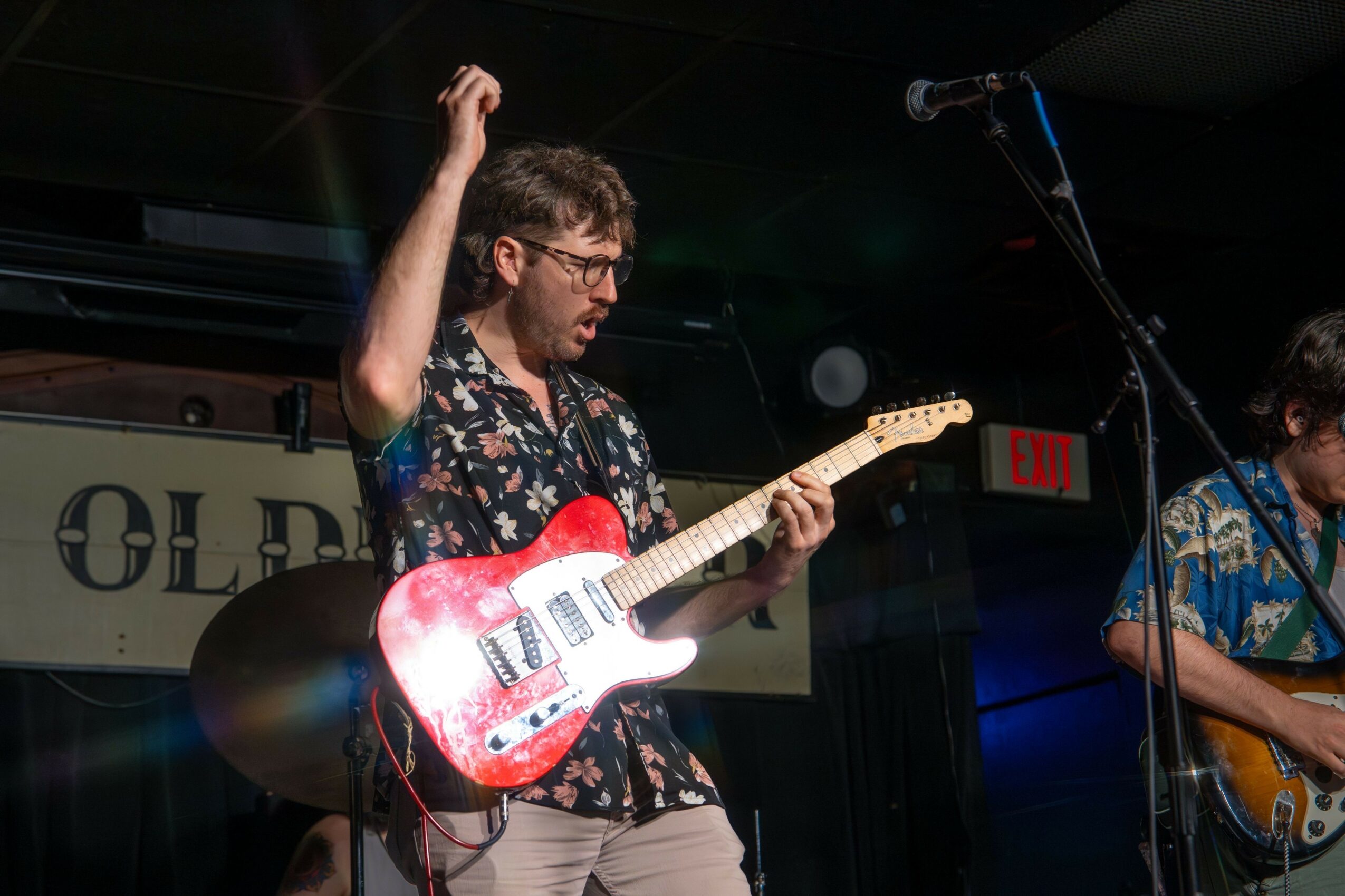 Man playing a red electric guitar on stage with a dark background and microphone, wearing glasses and a patterned shirt.