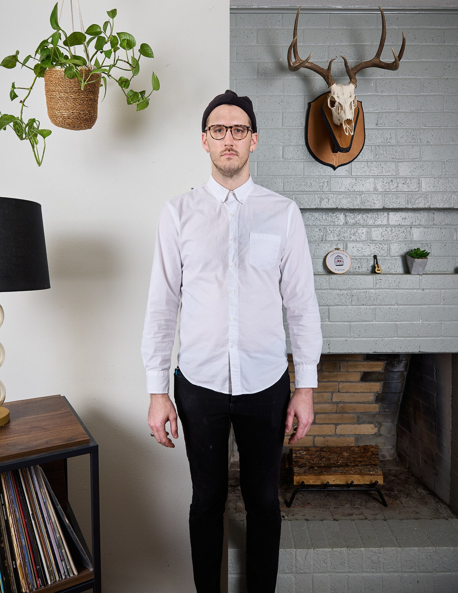Man with glasses in white shirt and black pants standing indoors, fireplace with mounted deer skull and plant on wall.