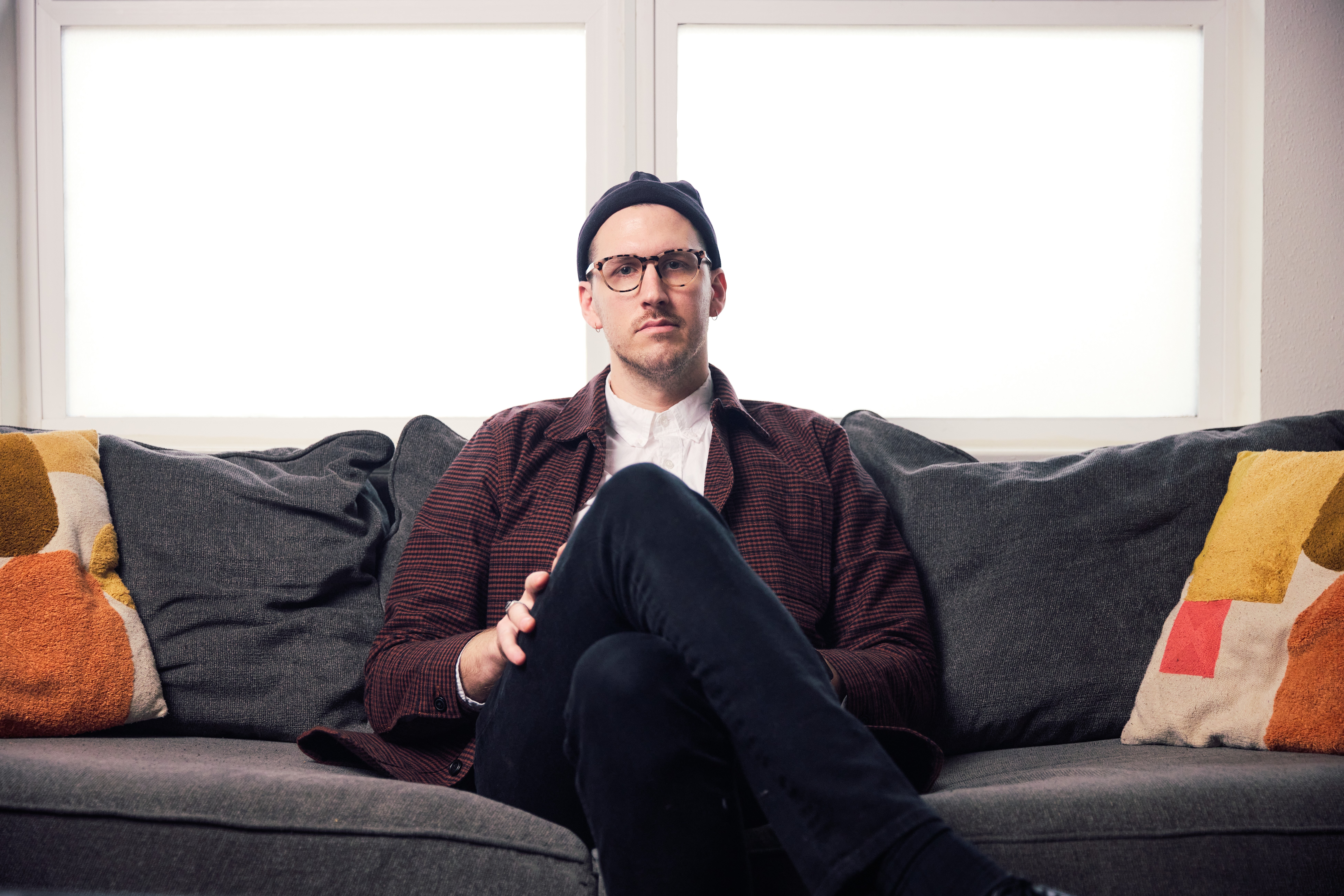 Man sitting on a sofa with crossed legs, wearing glasses, a dark jacket, and a white shirt, in front of large windows.