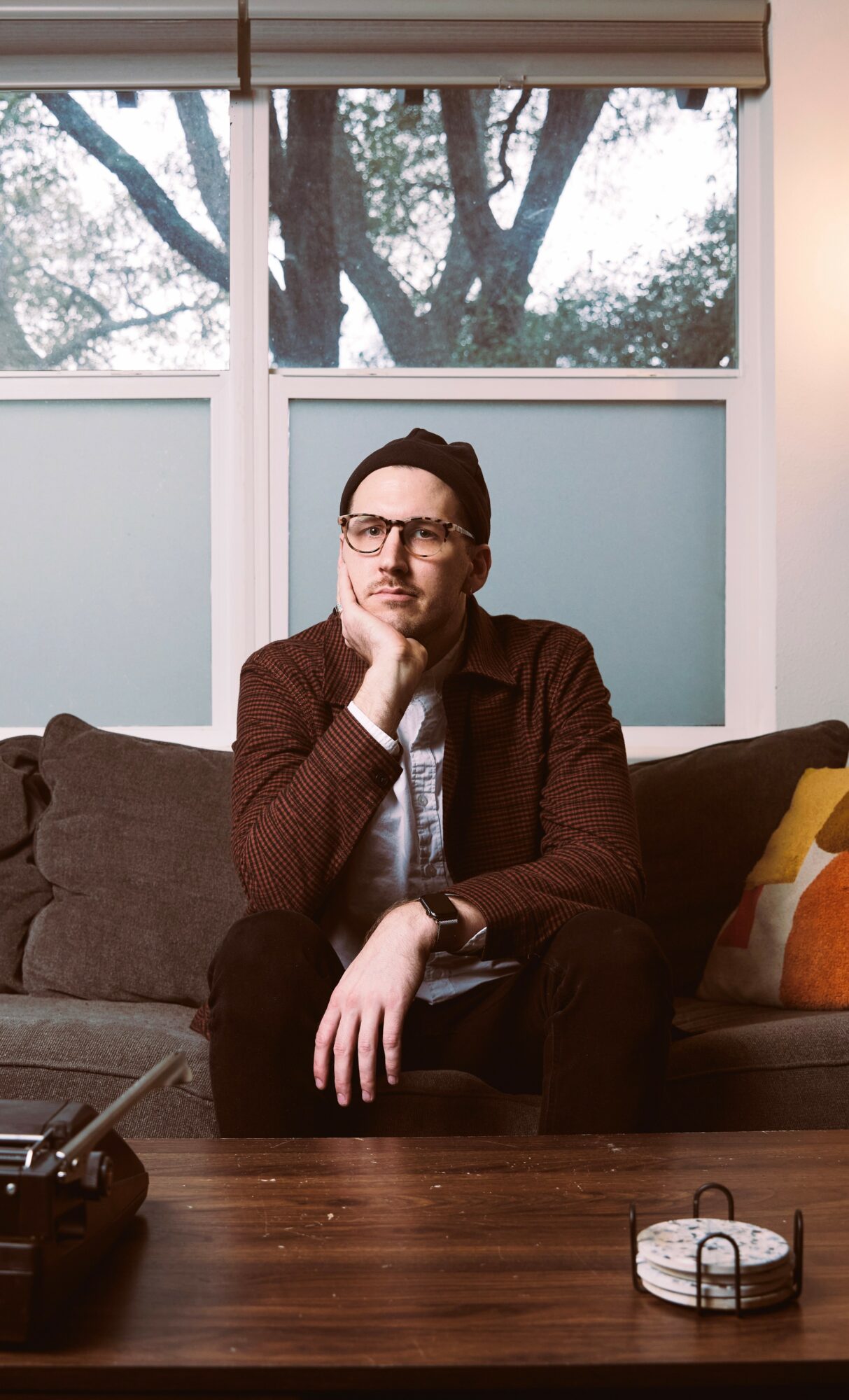 Man with glasses sitting on a sofa, resting his chin on his hand, in a room with large windows and trees outside.