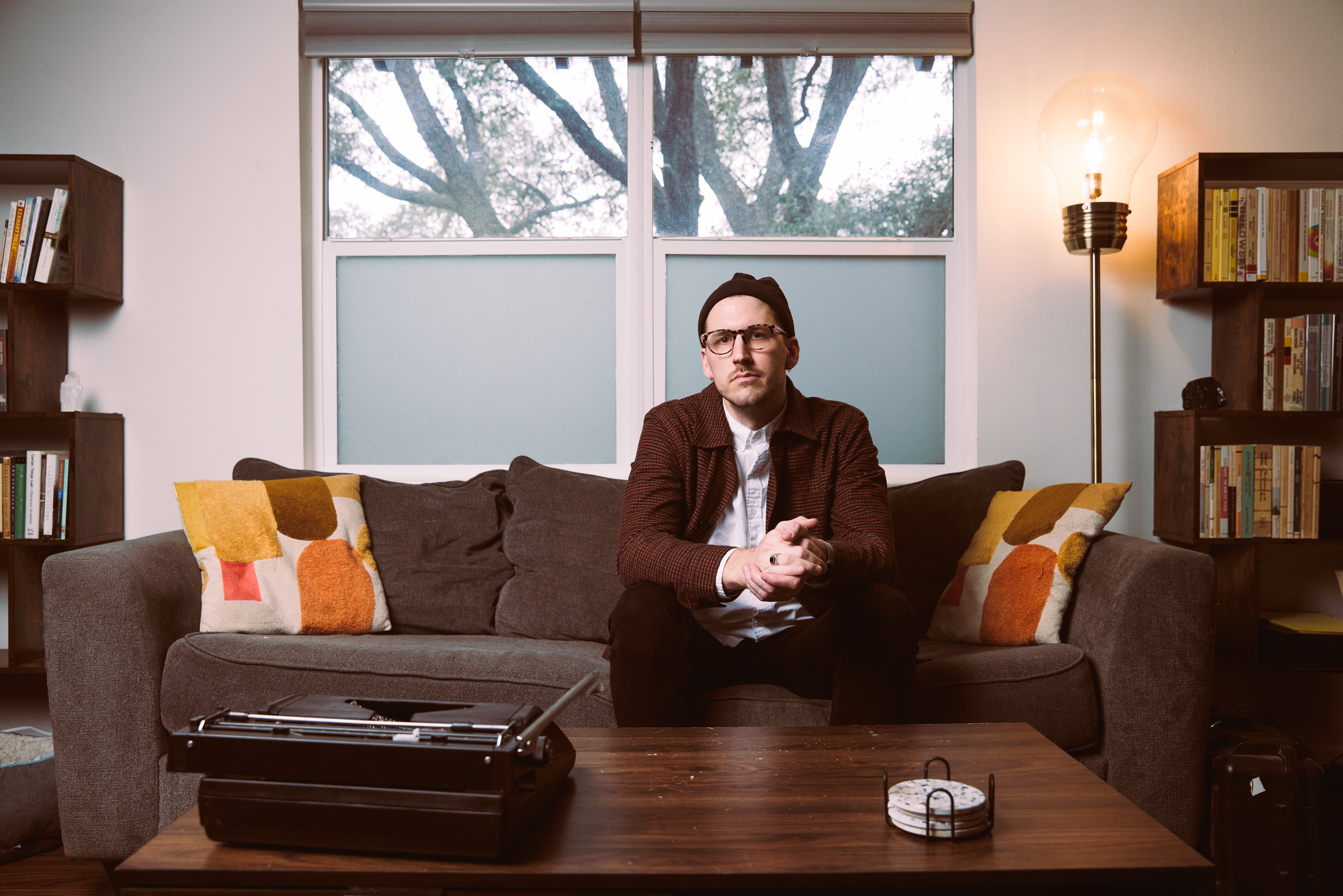 Man sitting on a sofa in a living room with bookshelves, window, and a coffee table in front.