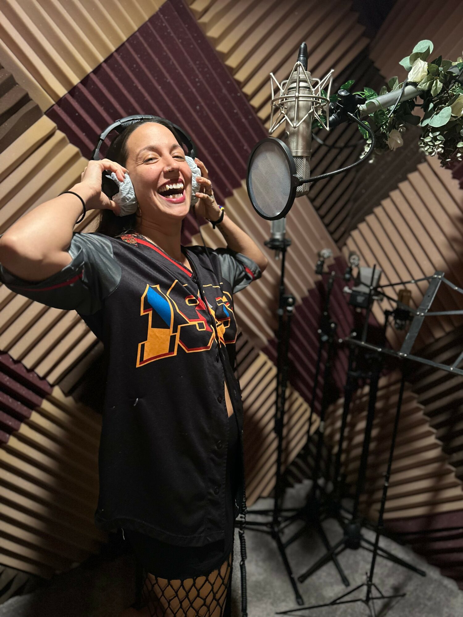 Young woman singing in a recording studio with a microphone and soundproof walls, smiling with headphones on.