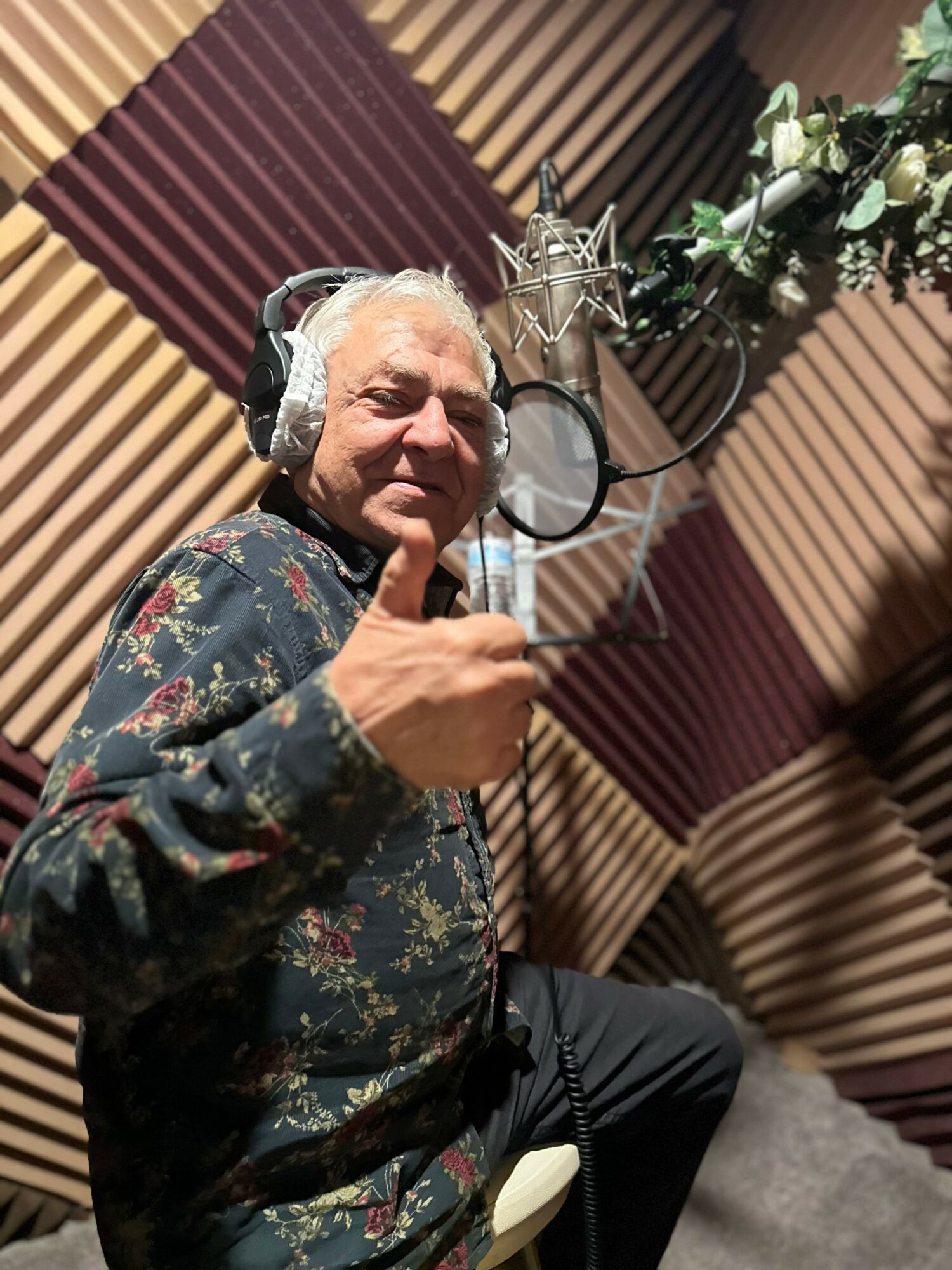 Older man with headphones in a recording studio, giving a thumbs-up gesture, with a microphone and floral decoration nearby.