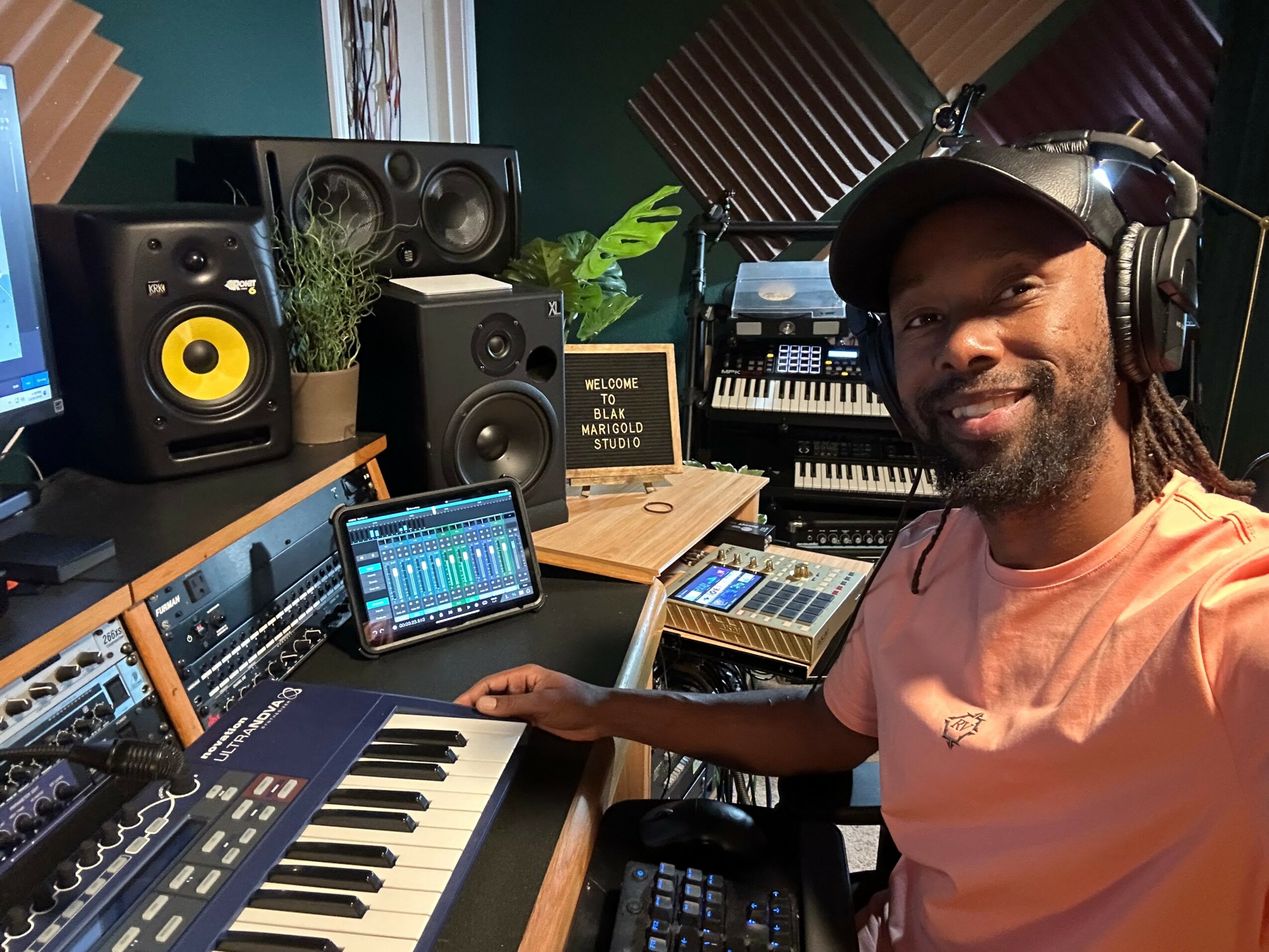Man in headphones smiling in a music studio with keyboards, speakers, and audio equipment.