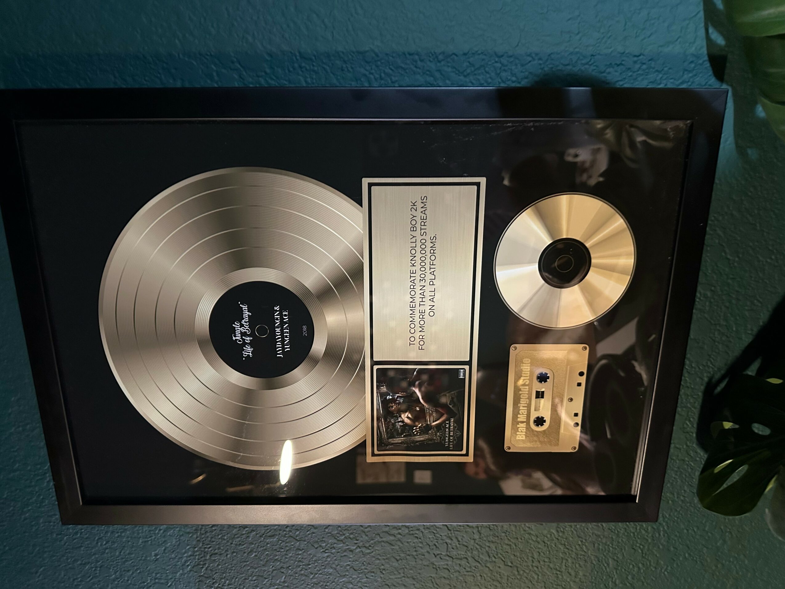 Framed gold record, a small photo, and a gold cassette tape displayed on a wall.