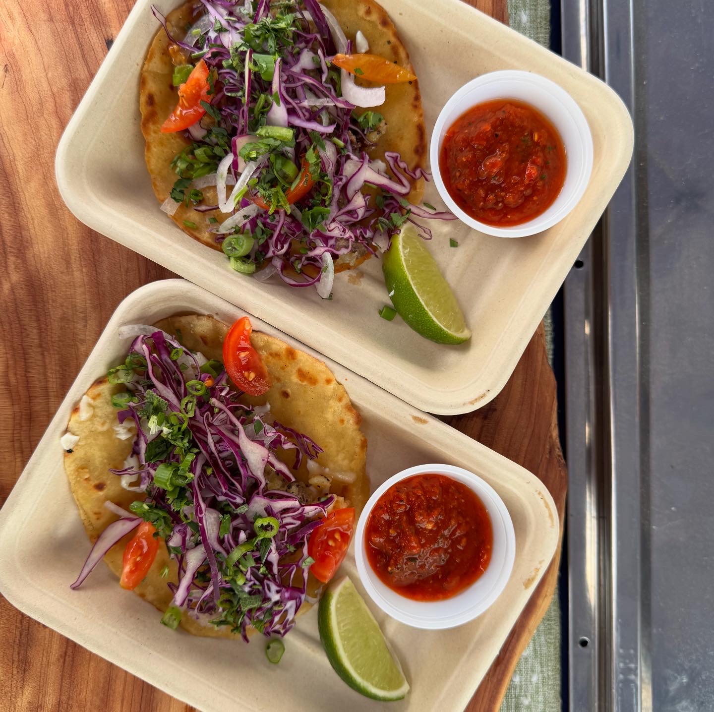 Two plates of tacos with purple cabbage, cherry tomatoes, lime wedges, and salsa, on a wooden table.