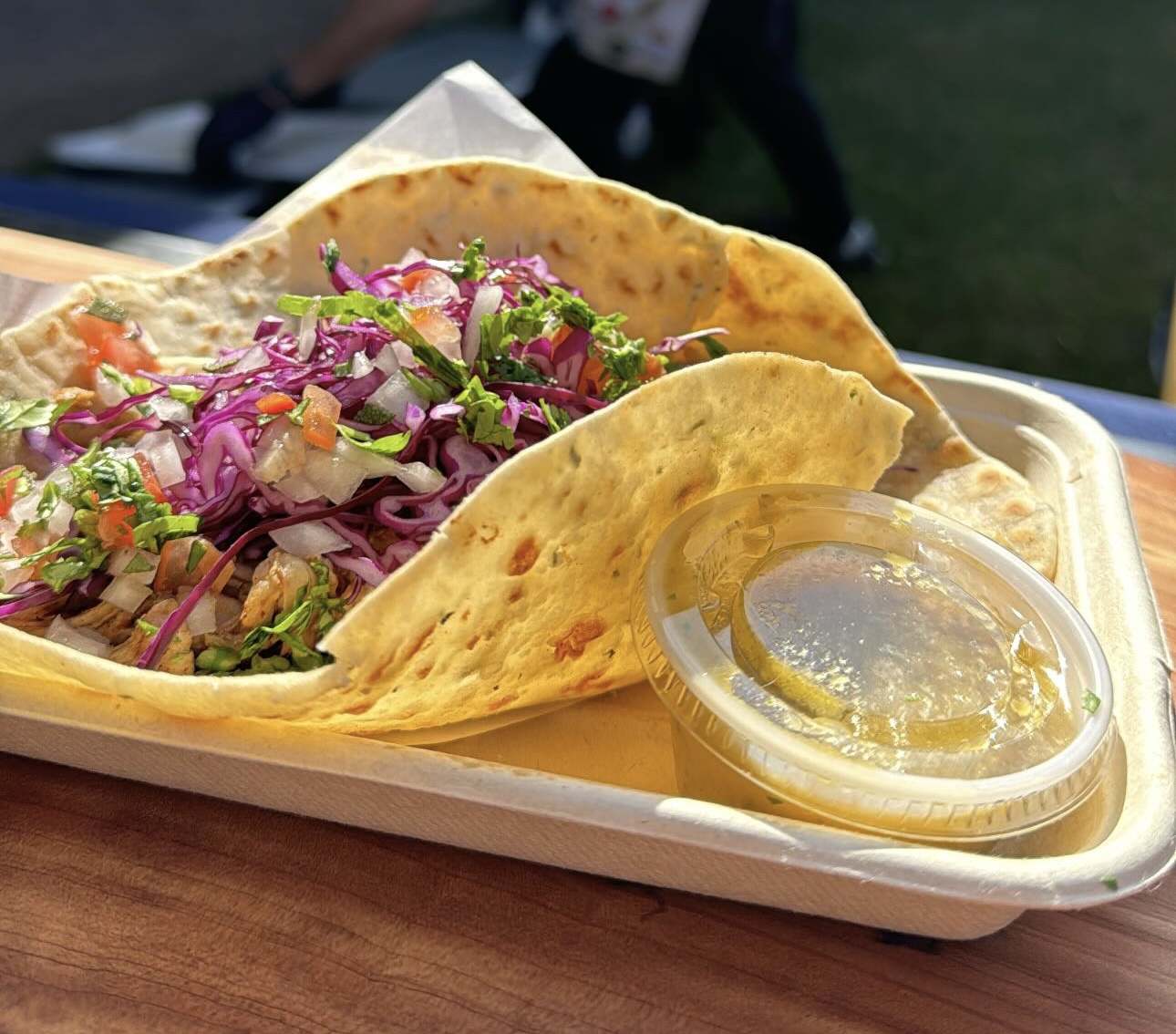 Two tacos filled with meat, purple cabbage, and vegetables, served with a small cup of sauce or dressing, on a tray.