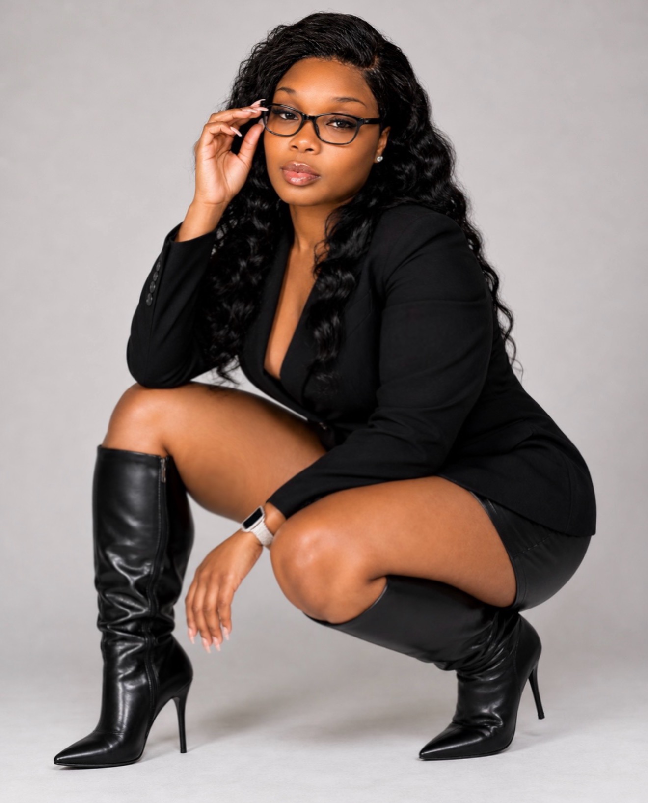 Woman crouching, wearing glasses, black blazer, and high-heeled boots, adjusting glasses with her right hand, against a plain background.