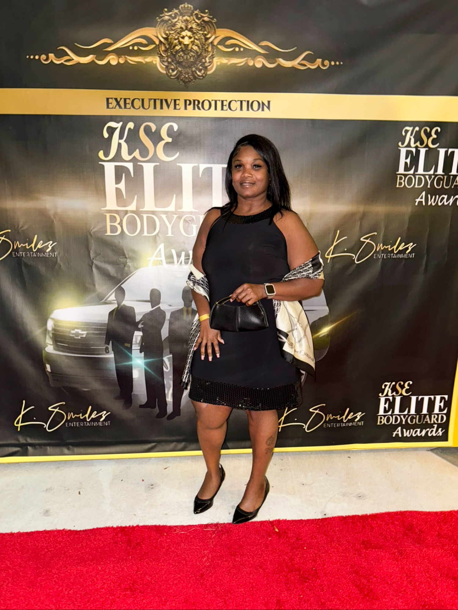 Woman in black dress and heels standing on red carpet in front of a black and gold backdrop with event text.