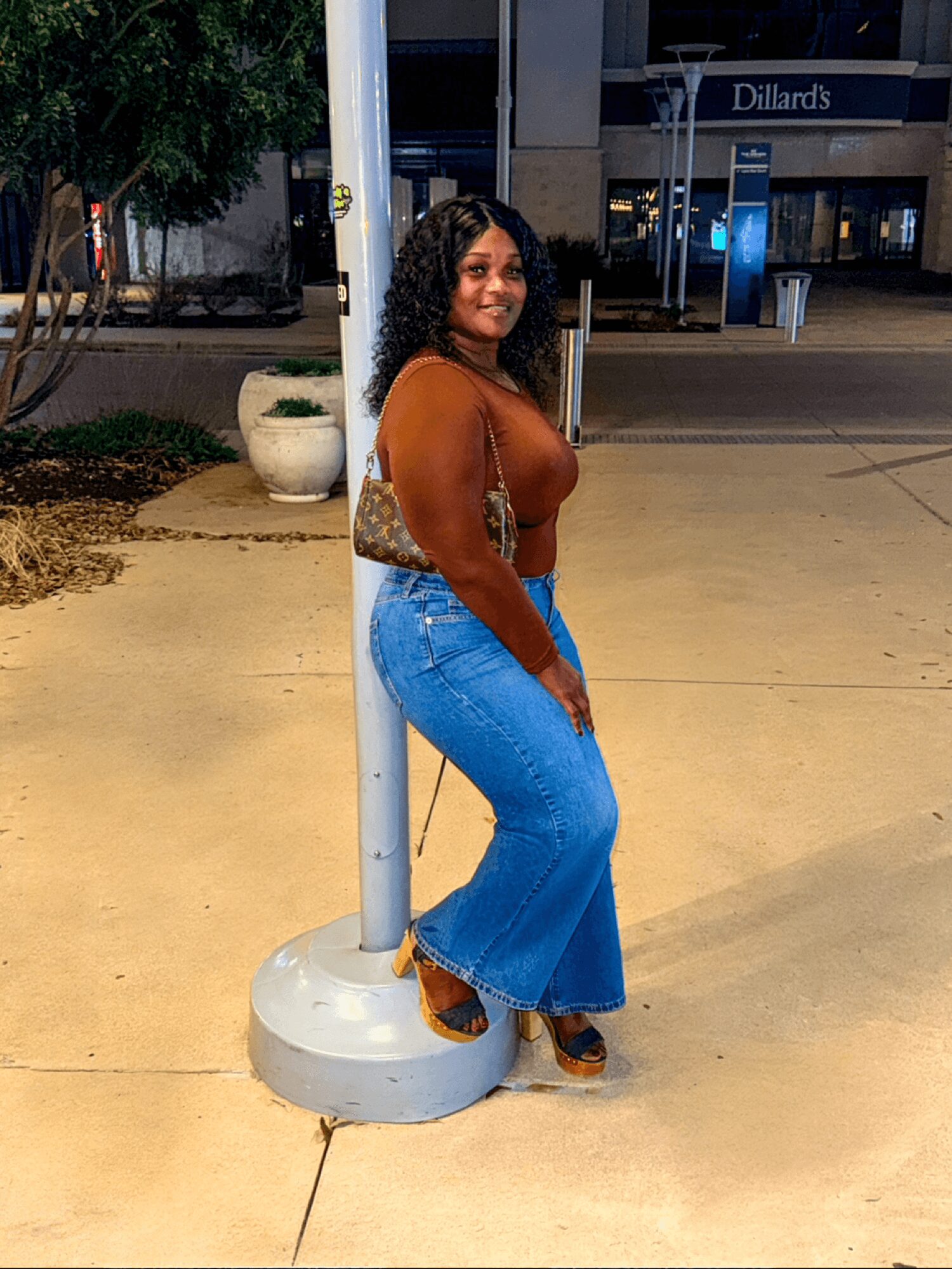 Woman standing outdoors at night, leaning against a pole, smiling, wearing a brown top and blue jeans.