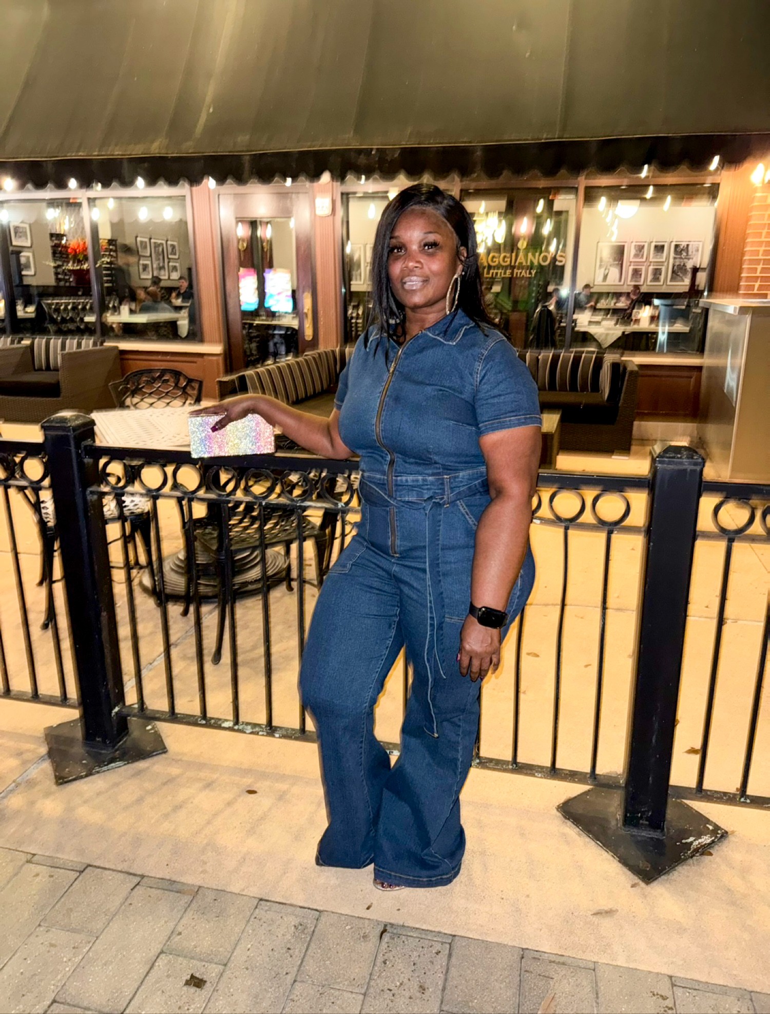 Woman in denim outfit standing outdoors near a black fence, holding a paper, with a restaurant in the background.