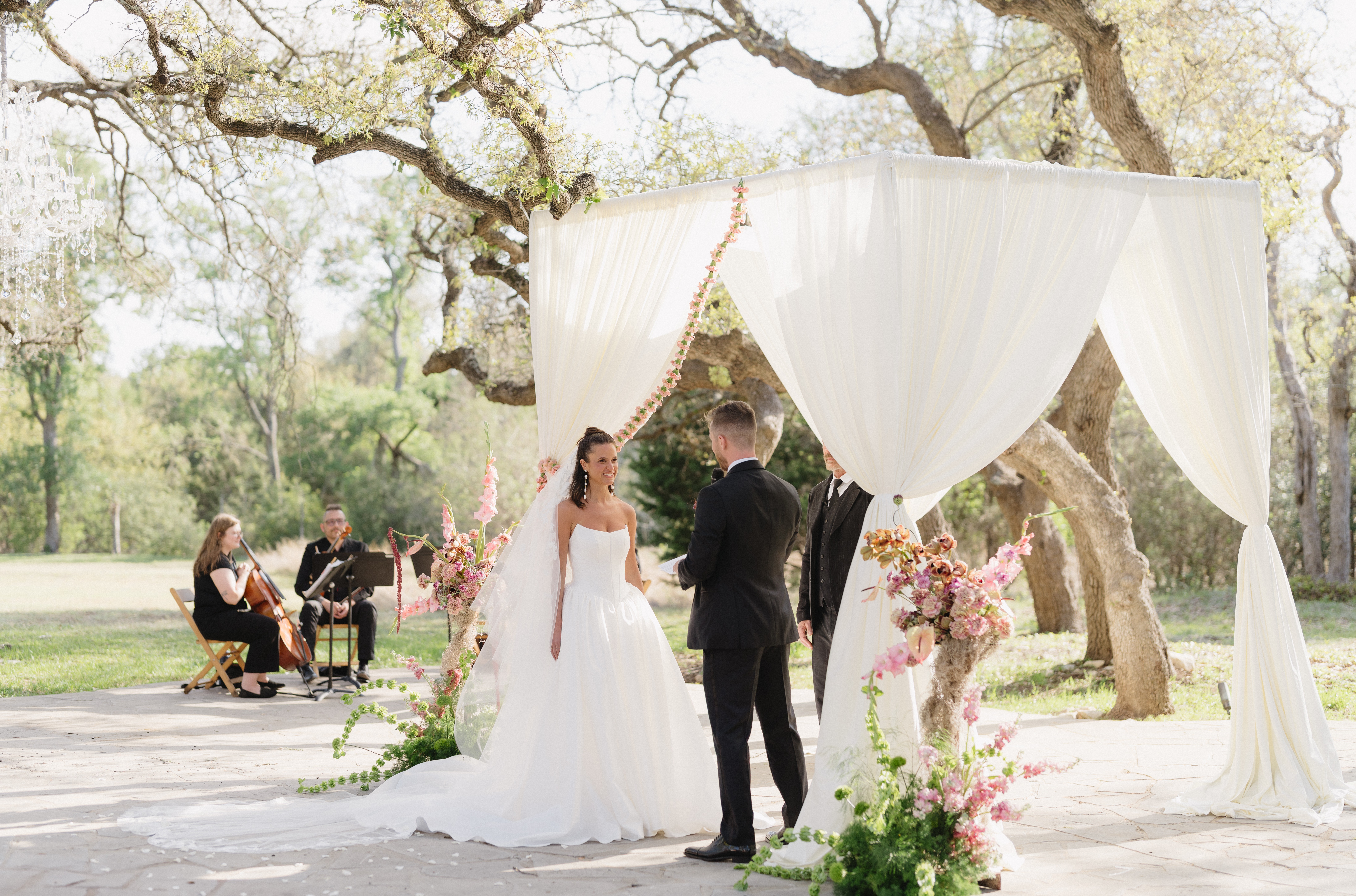 Bride and groom standing under a white canopy during outdoor wedding ceremony, with musicians playing in background, trees and sunlight.