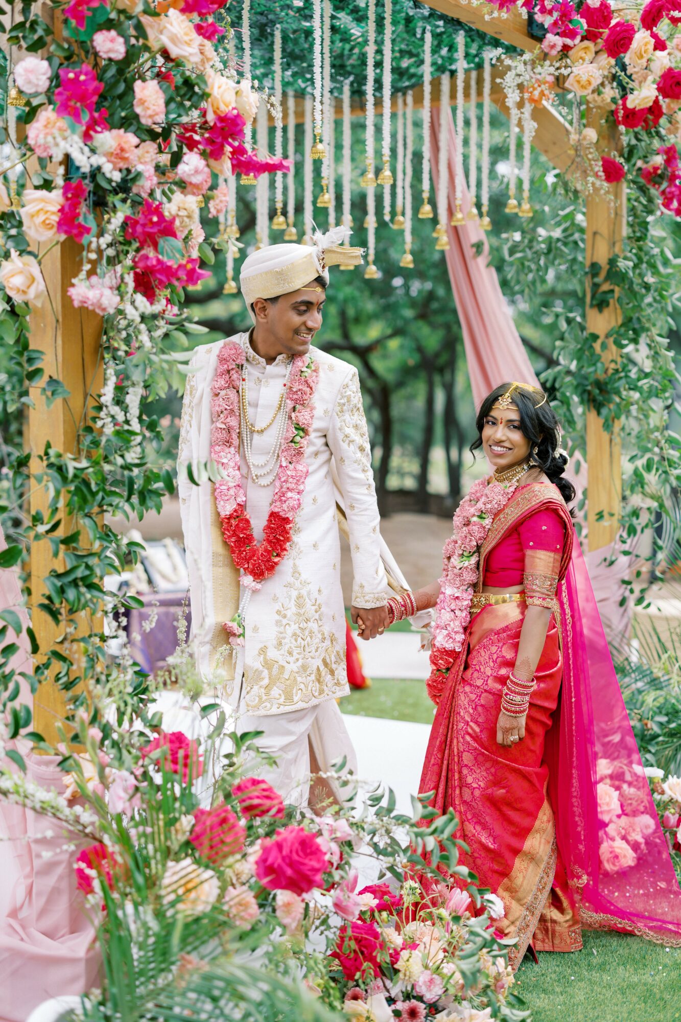 A couple in traditional attire holding hands at a decorated outdoor wedding ceremony with flowers and drapes.