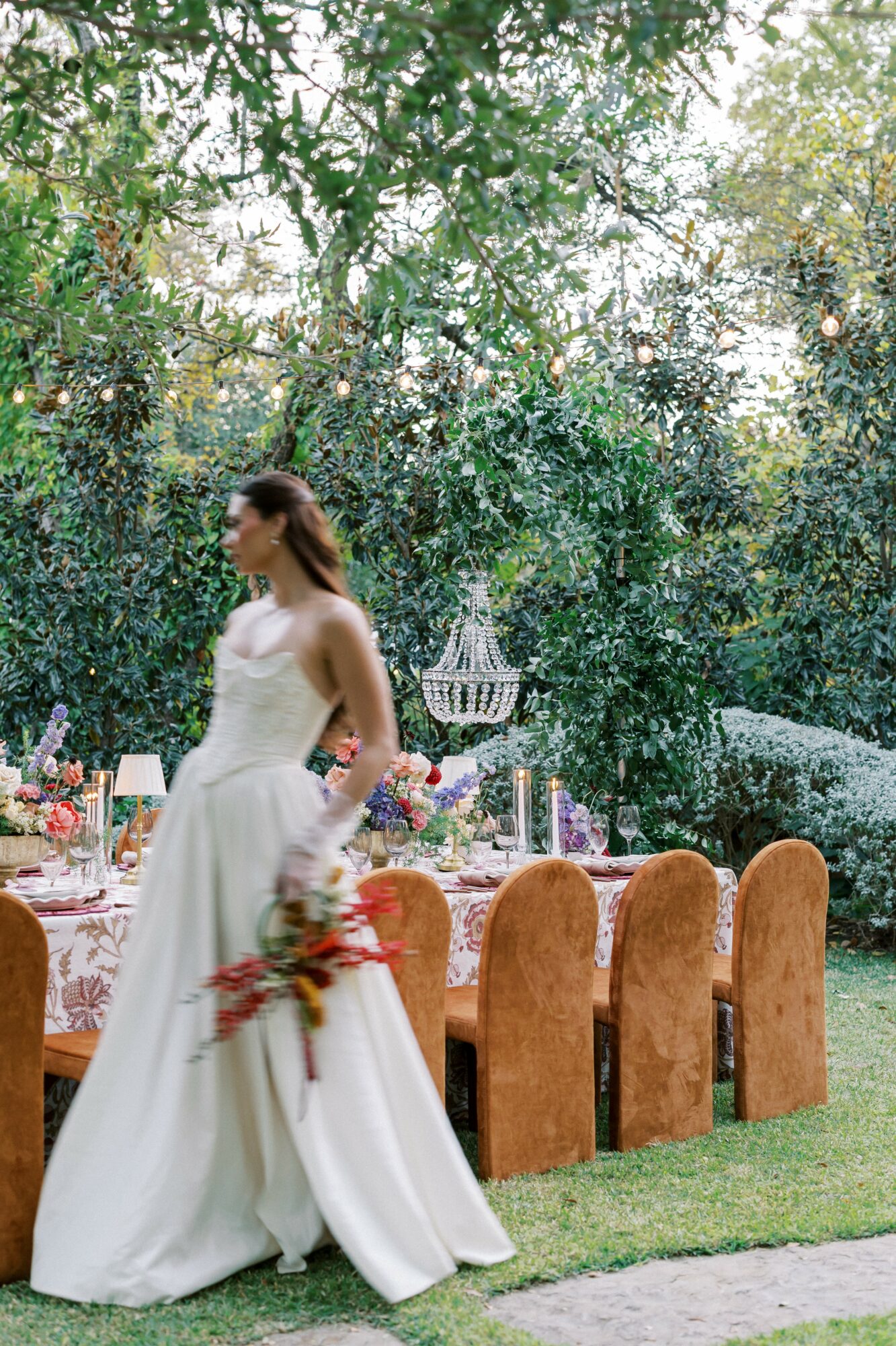 Woman in wedding dress holding flowers outdoors near a decorated table with chairs, surrounded by trees and greenery.