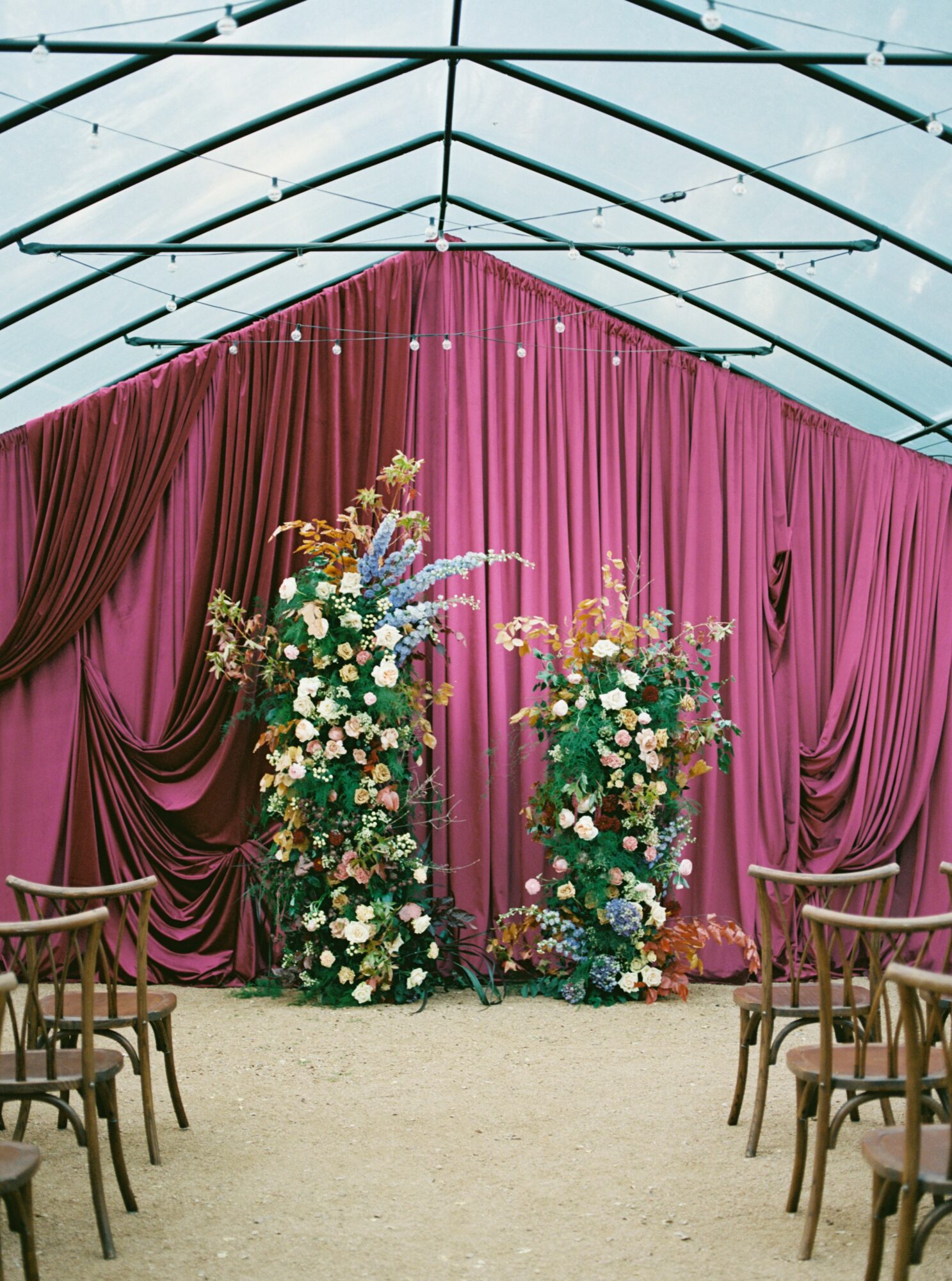 Indoor event space with pink curtains, floral arrangements, and chairs arranged on both sides, under a glass ceiling.