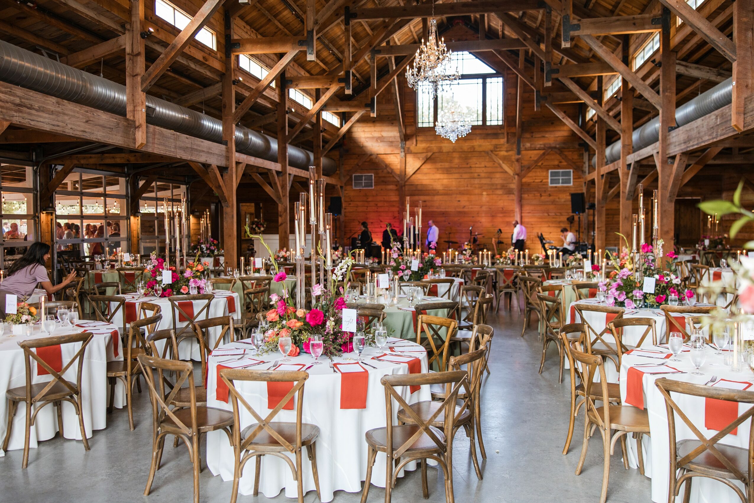 Elegant event space with round tables, floral centerpieces, and wooden beams, decorated for a celebration or wedding reception.