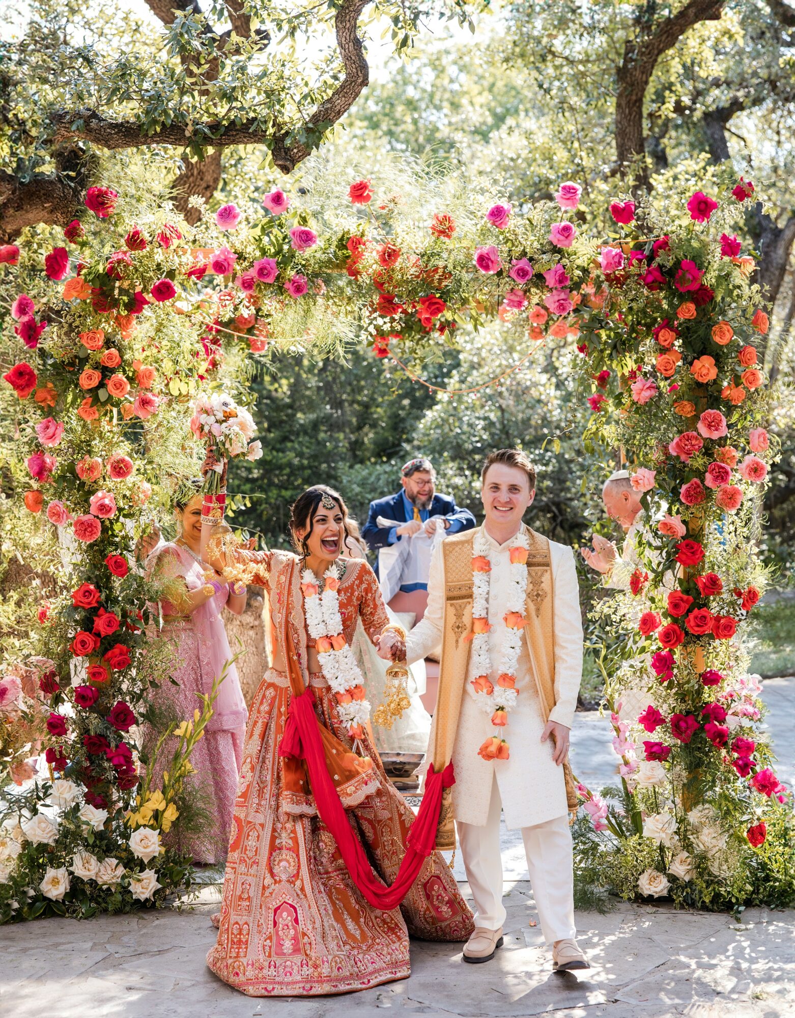 A wedding ceremony outdoors with a floral arch, bride and groom, and guests, under trees with sunlight filtering through.