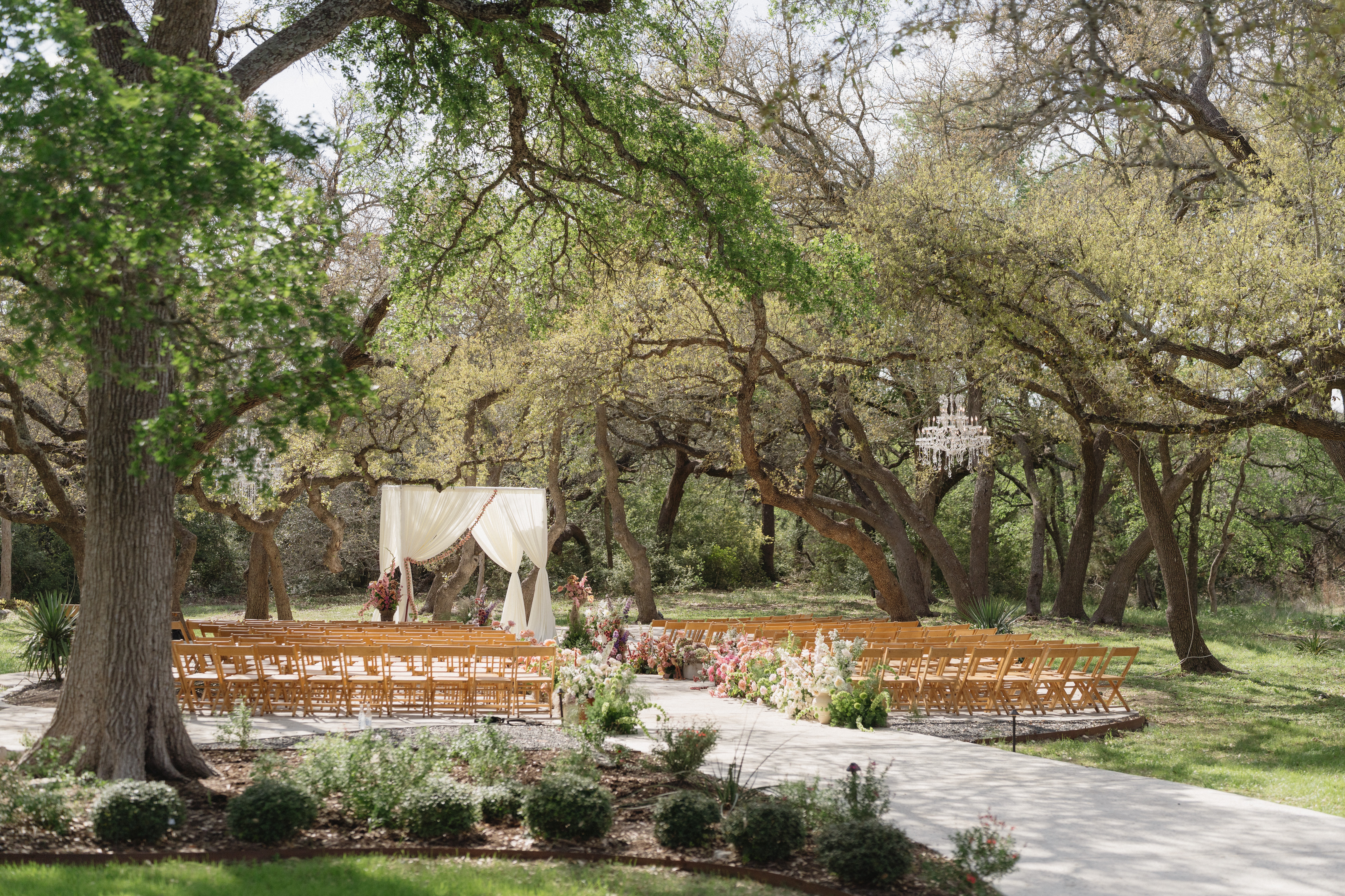 Outdoor wedding setup with chairs, trees, and a white draped arch in a park-like setting.