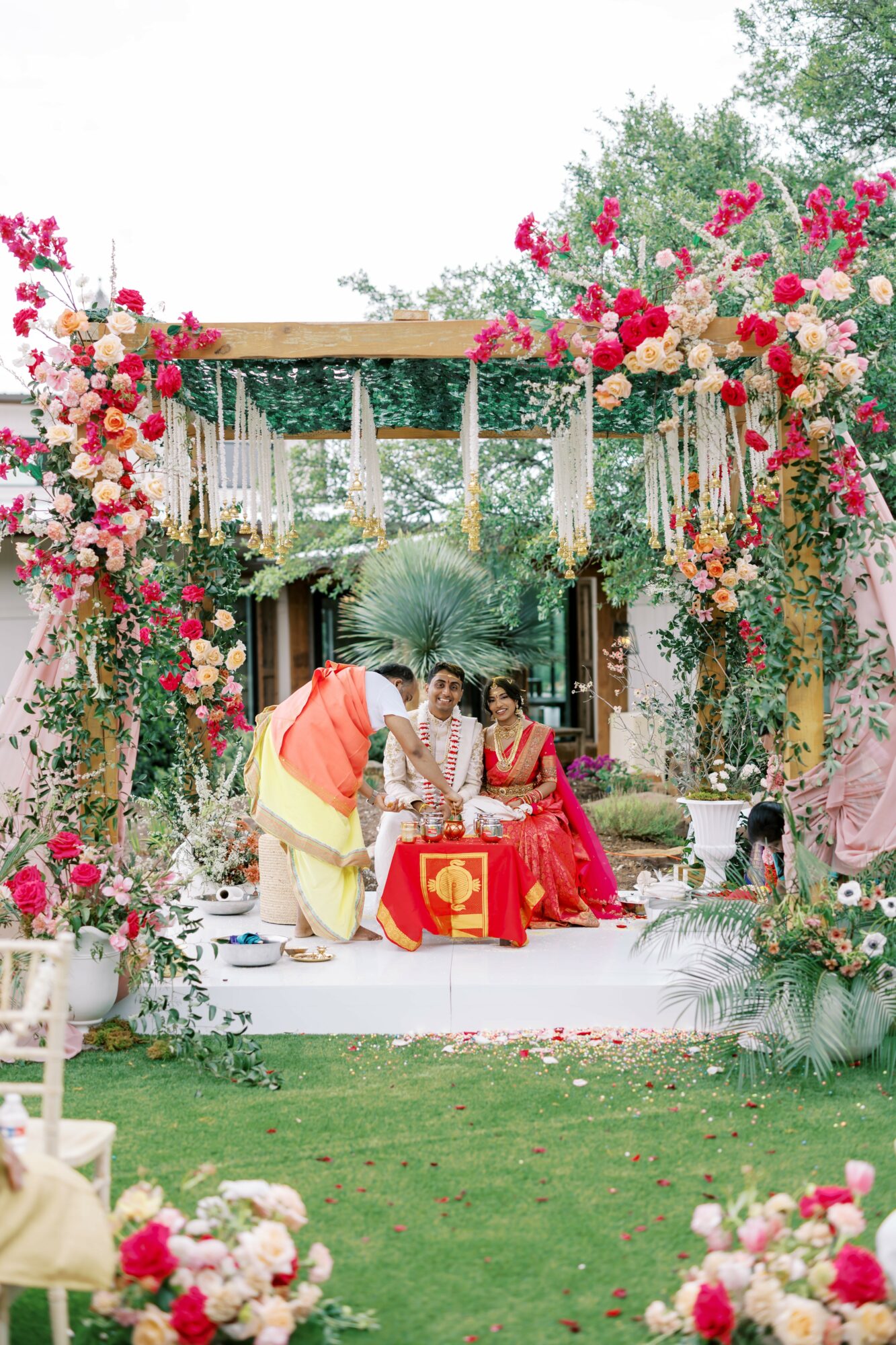 Three women sitting on a decorated stage with floral arrangements and greenery, outdoors during daytime, celebrating a special event.