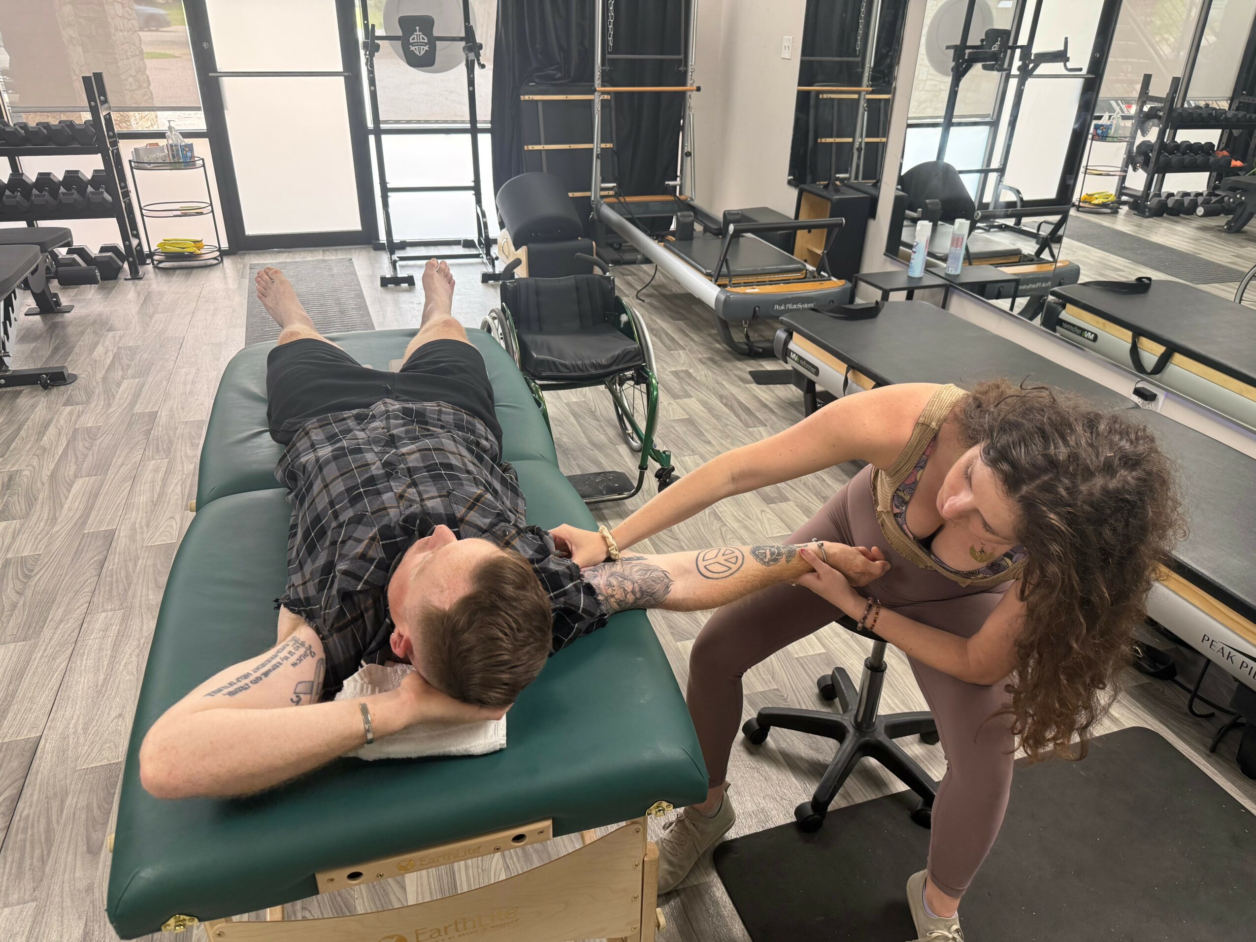 Tattooed woman with curly hair leaning over a person lying face down on a massage table in a gym or therapy room.