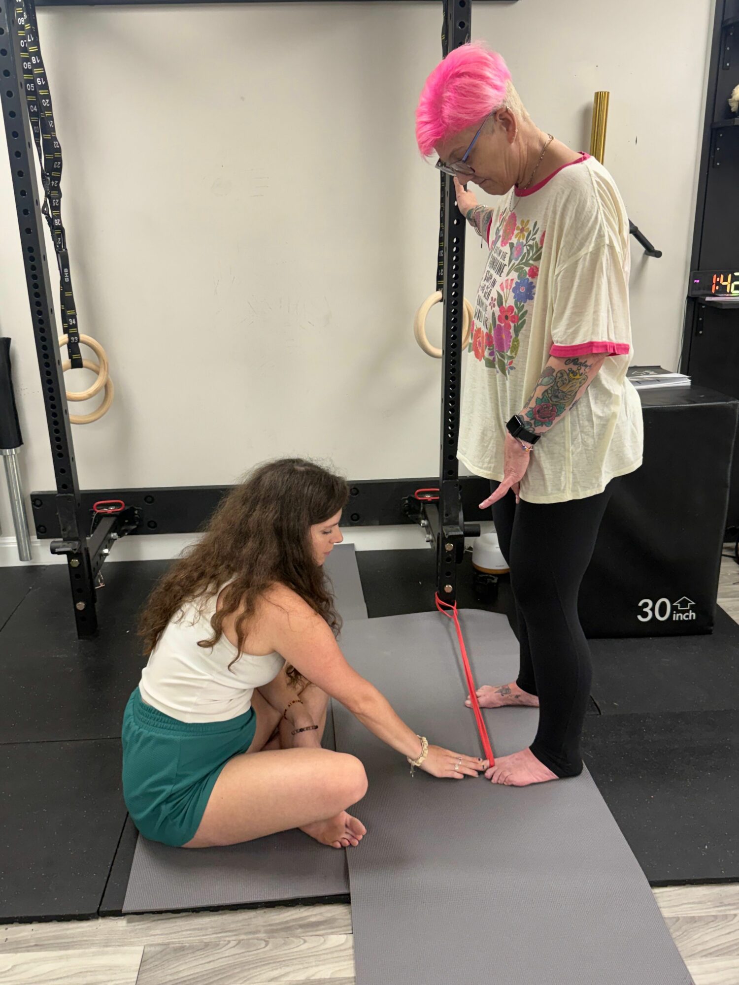 Person with pink hair standing in gym, another person sitting on the floor stretching, gym equipment in background.