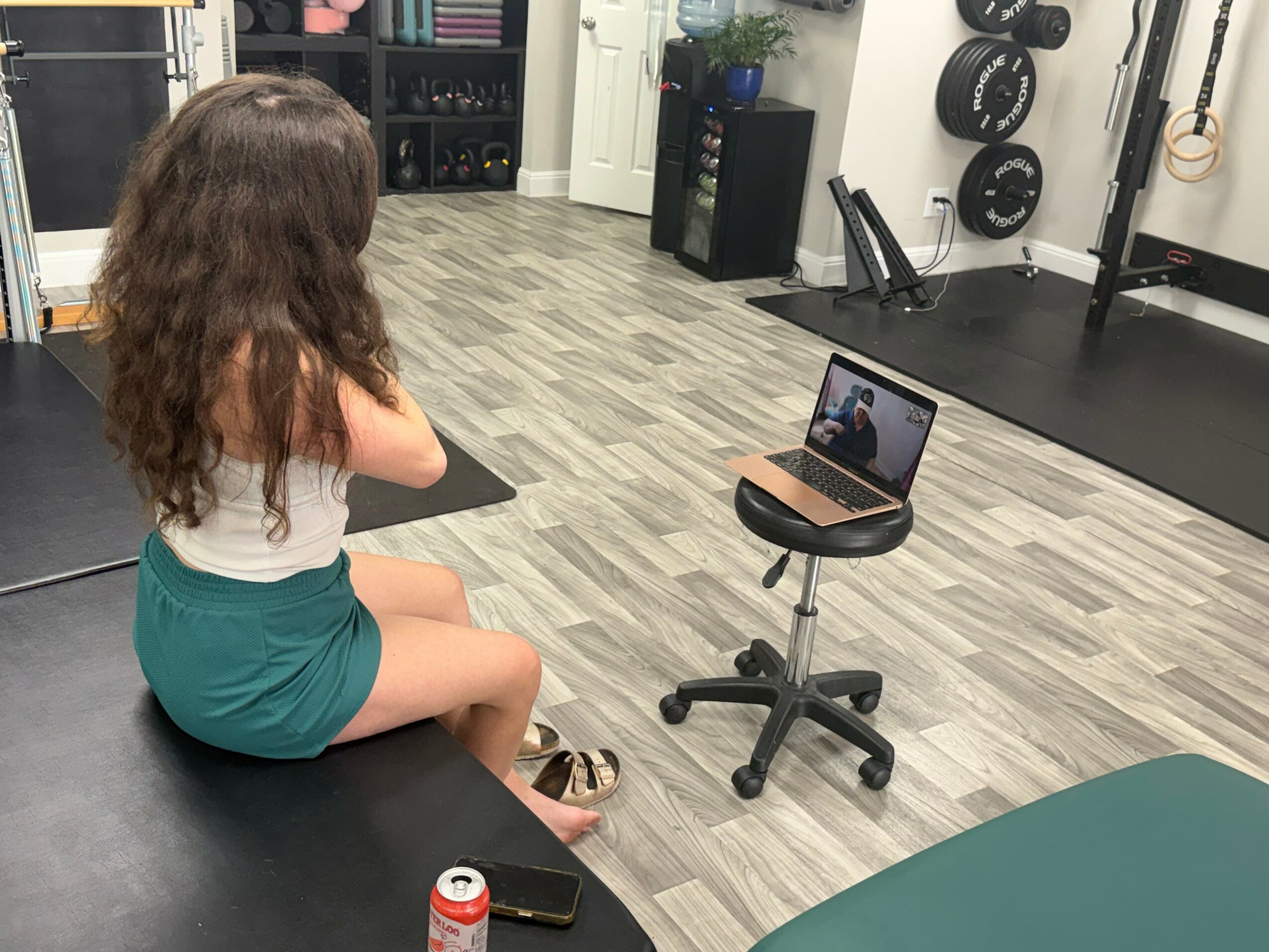 Girl sitting on a bench in a gym, looking at a laptop on a stool, with gym equipment in the background.