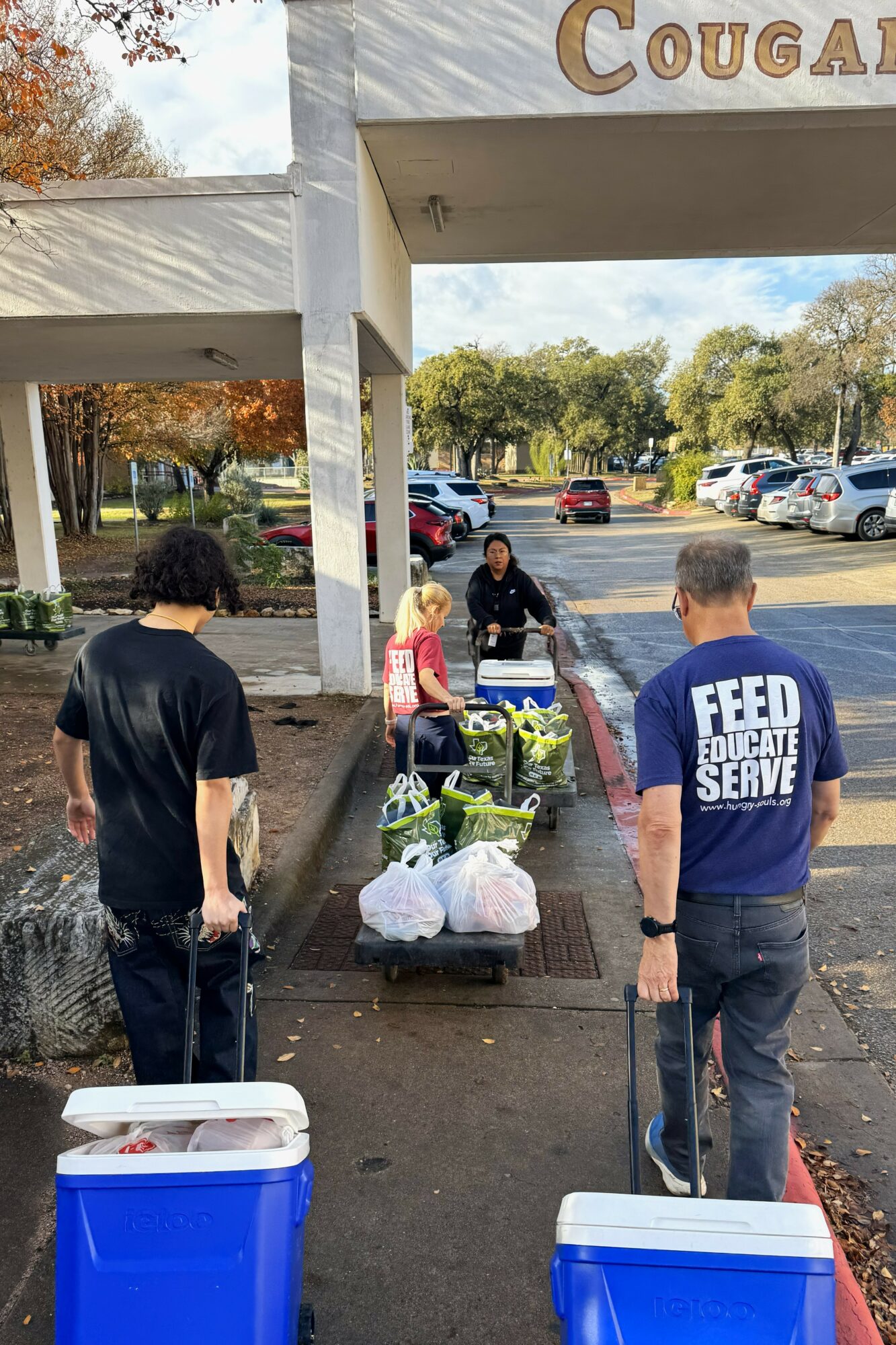 People gather outside a building with a sign reading 'Cougar,' with carts and supplies nearby, trees in the background.