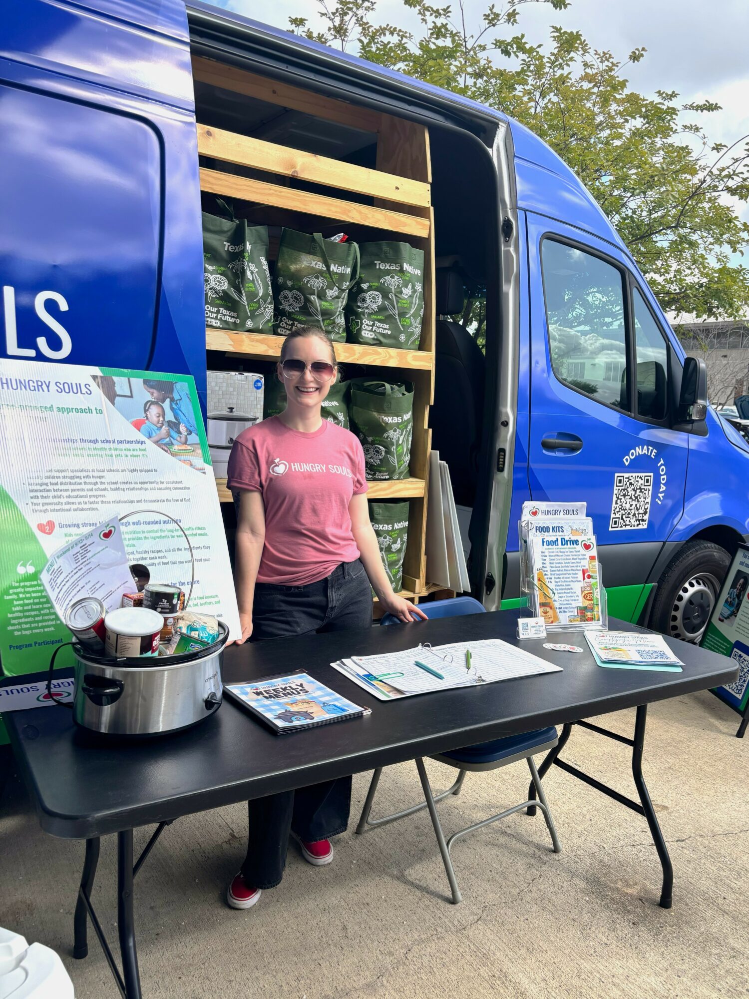 Woman standing behind a table with informational materials, next to a blue mobile unit with shelves of bags, outdoors.