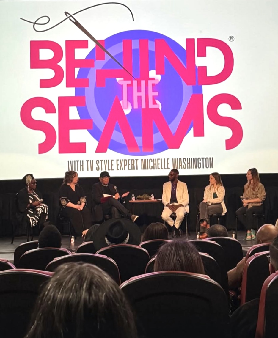 Panel discussion with five people seated on stage, audience in foreground, behind a large screen displaying the show's logo.