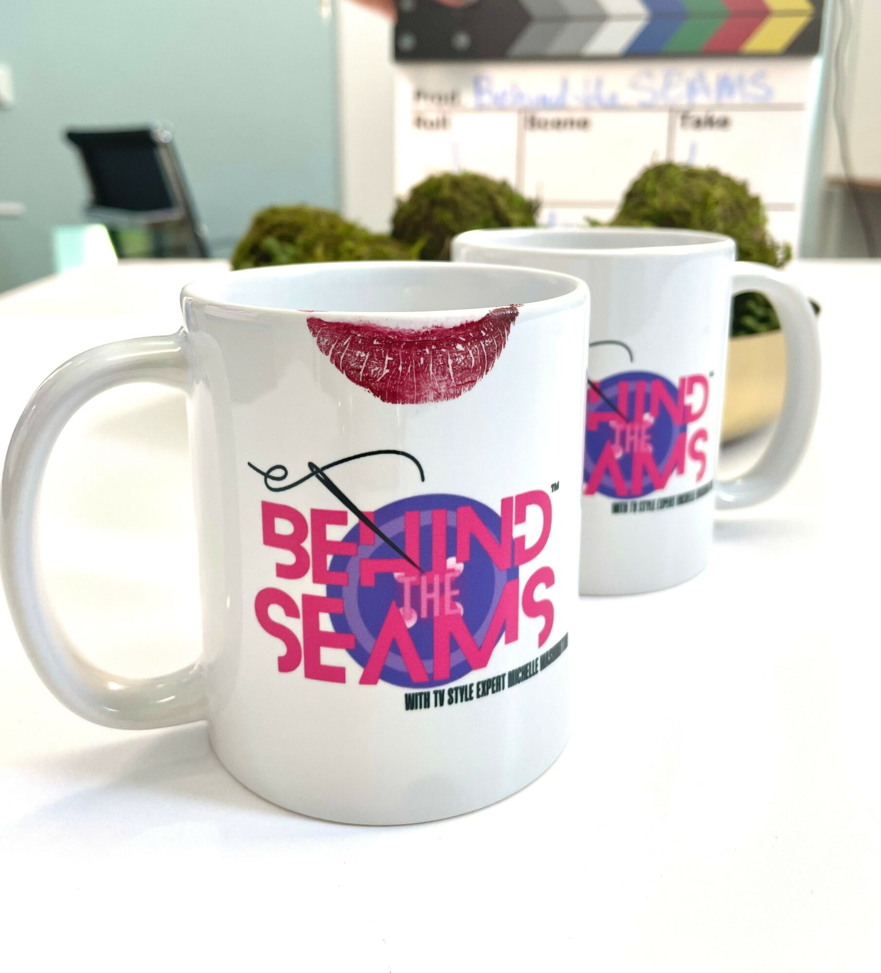 Two white mugs with colorful text and a lipstick mark, placed on a table with a blurred background.