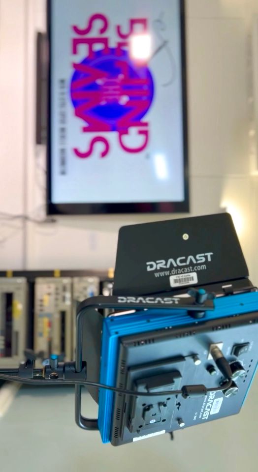 Close-up of a blue and black camera or projector equipment in front of a blurred television screen displaying pink and purple text.