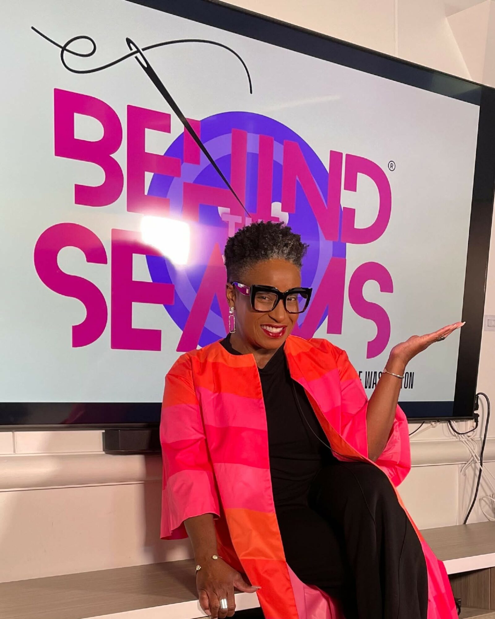 Woman with glasses and curly hair sitting in front of a screen displaying the words 'BEHIND SEAMS' with a needle and thread graphic, gesturing with her right hand.