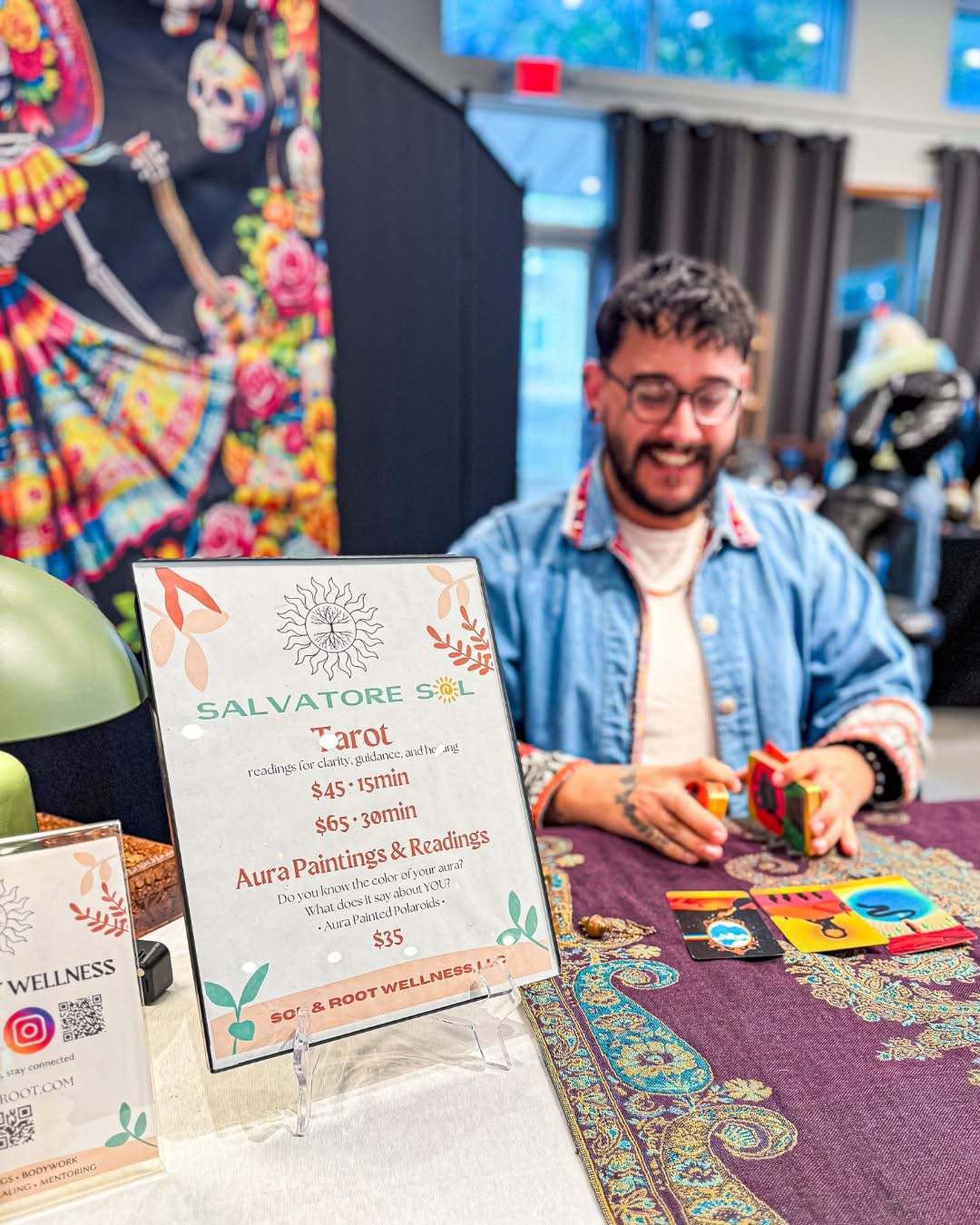 Man with glasses and beard smiling at a table with tarot cards, colorful background, and a sign about tarot readings.