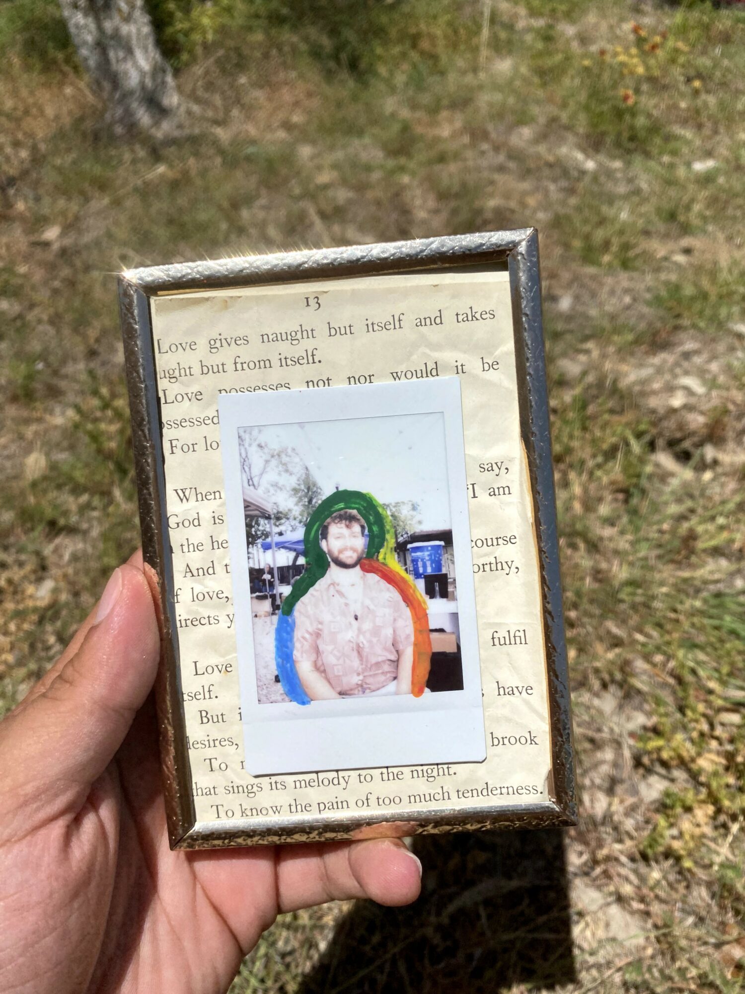 Person holding a framed photo outdoors, with a background of grass and trees, wearing a face mask and a pink top.