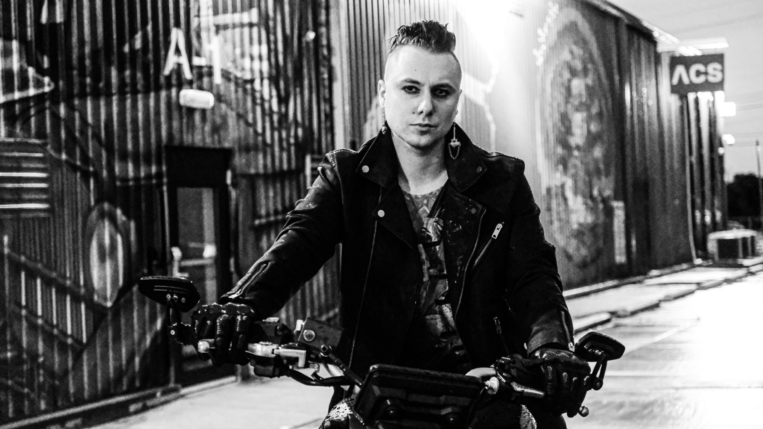 Person with short hair and leather jacket sitting on a motorcycle in an urban setting, black and white photo.