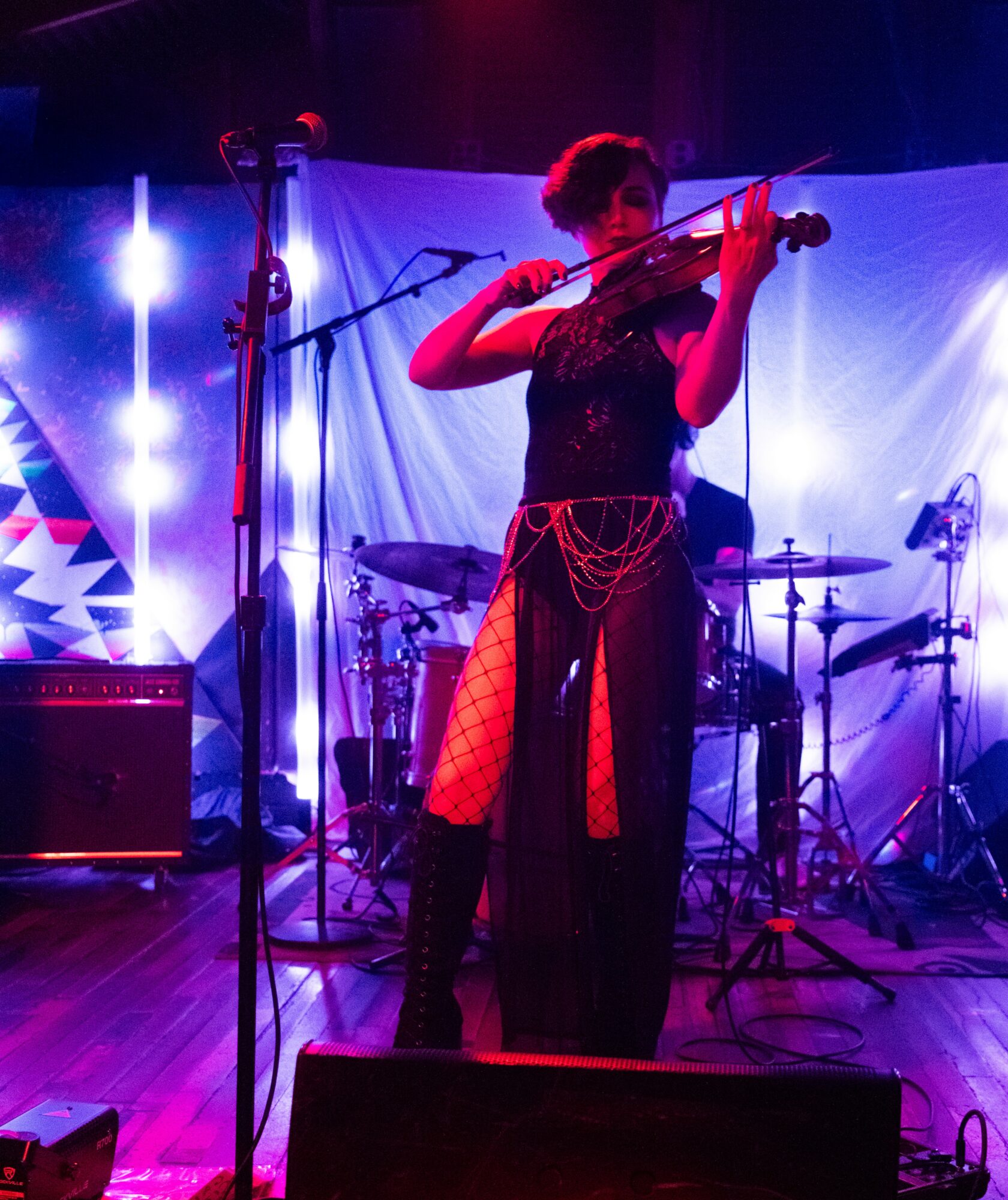 Person playing violin on stage with drums and colorful backdrop, stage lighting, and microphone stand.